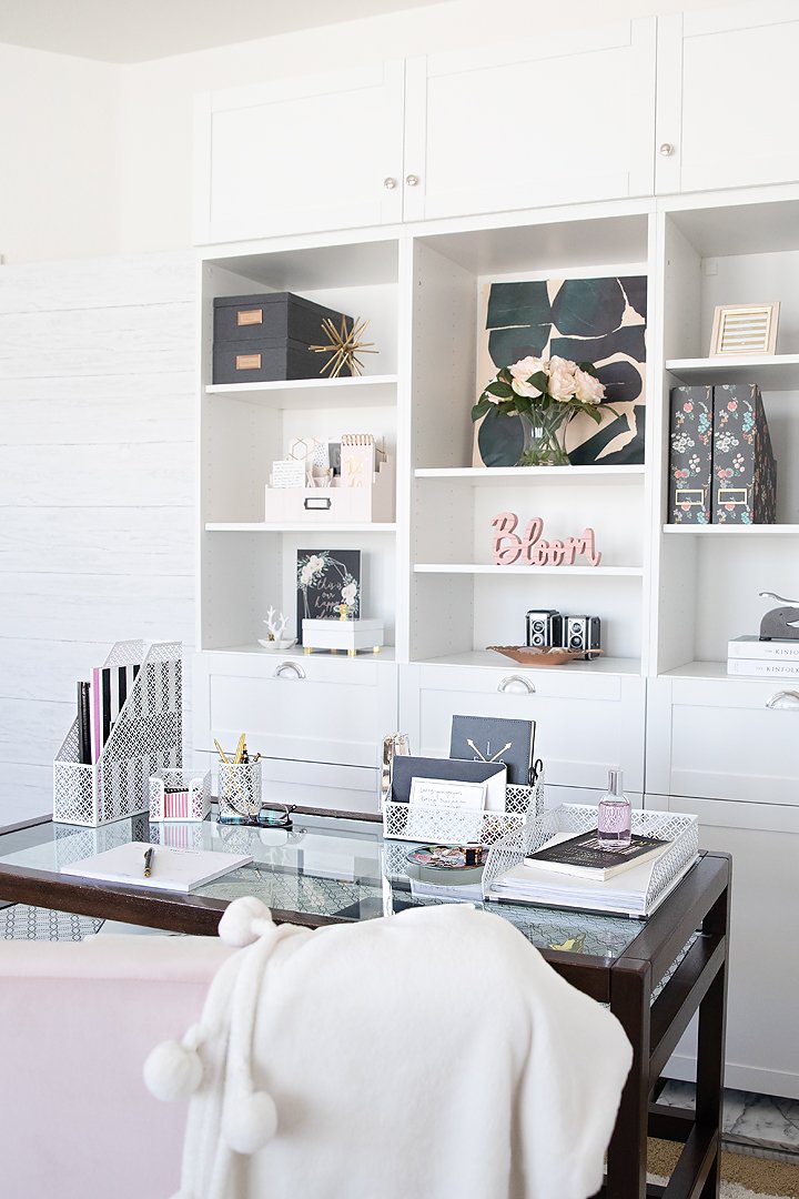 A bright home office features white shelving, a glass-top desk, and various cute, organized desk accessories.