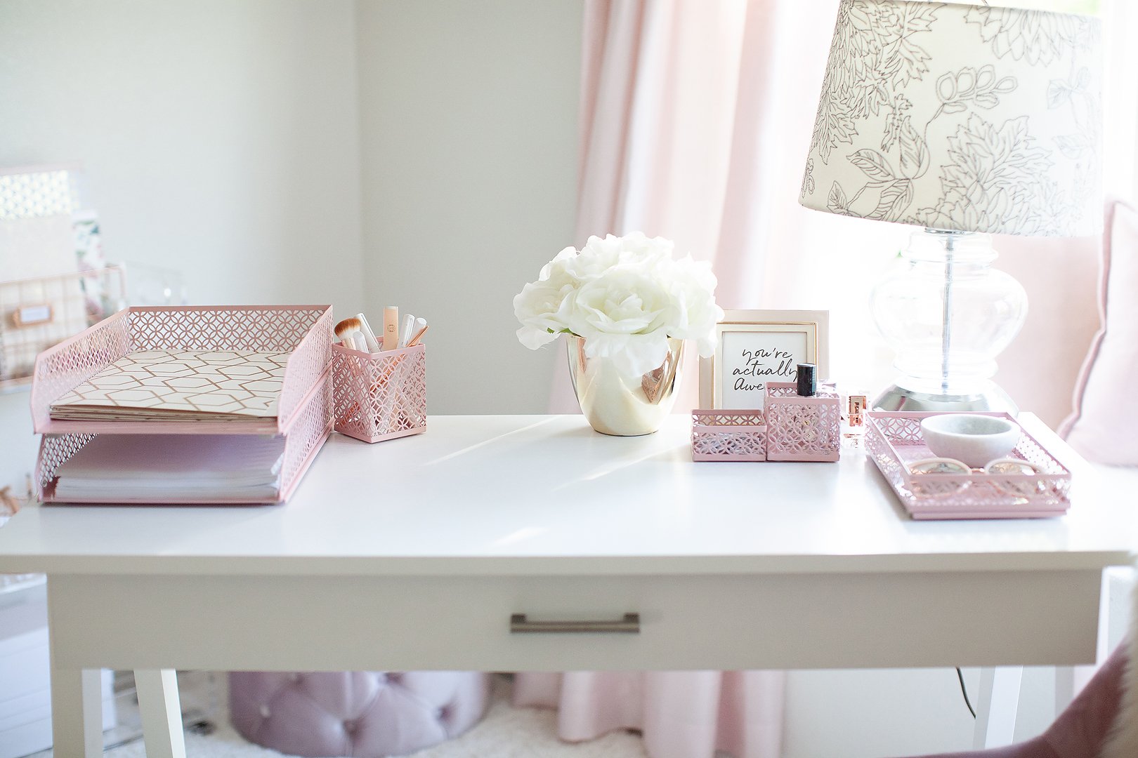 A beautifully organized white desk with pink office supplies, white flowers, and a decorative lamp, creating a soft aesthetic.