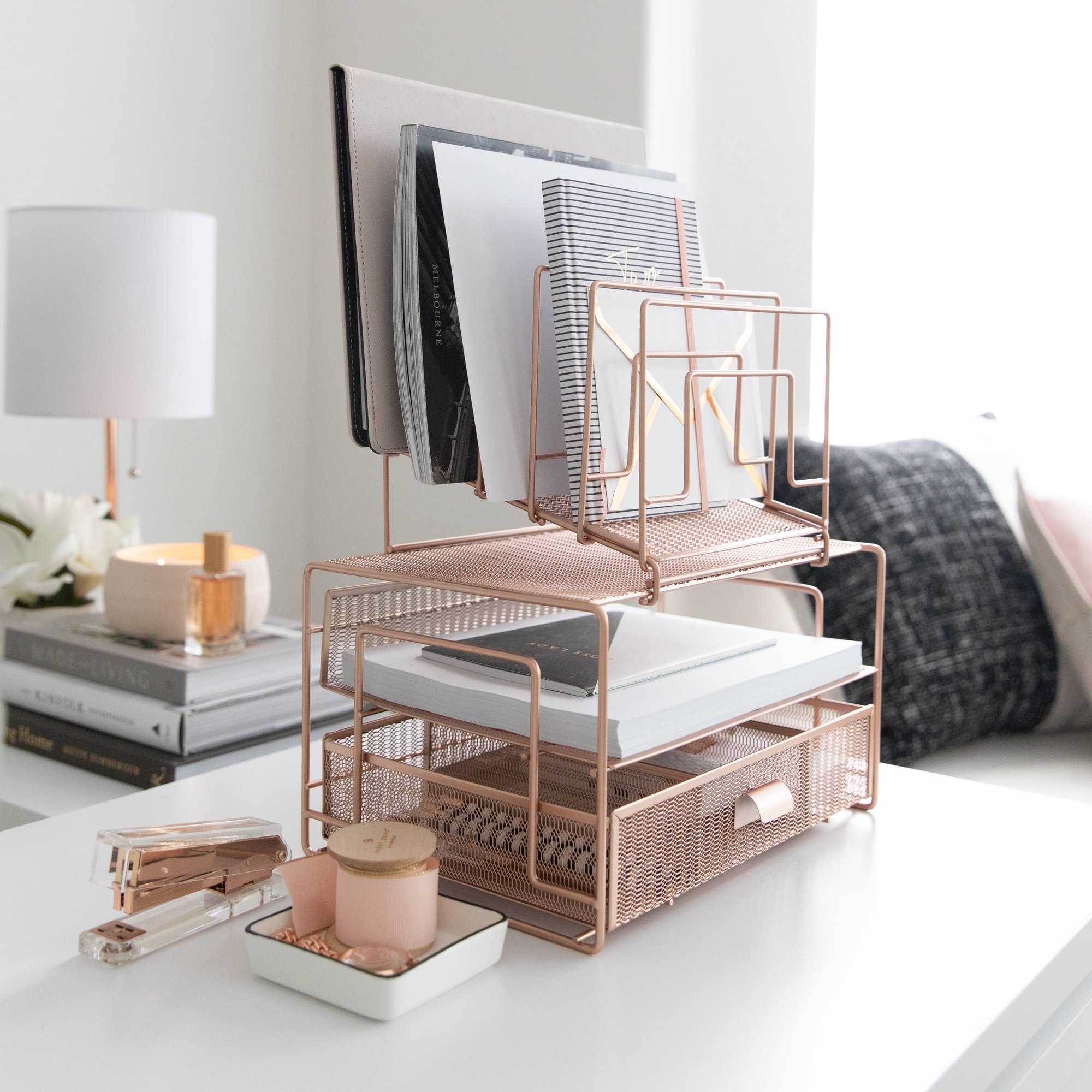A rose gold mesh desk organizer filled with notebooks and papers sits on a white desk.
