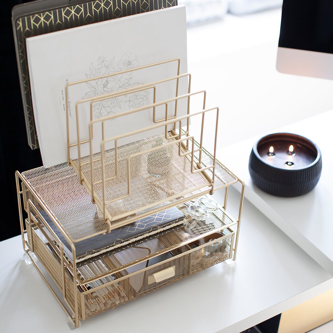 A stylish gold mesh desk organizer with papers, notebooks, and a candle on a white desk.