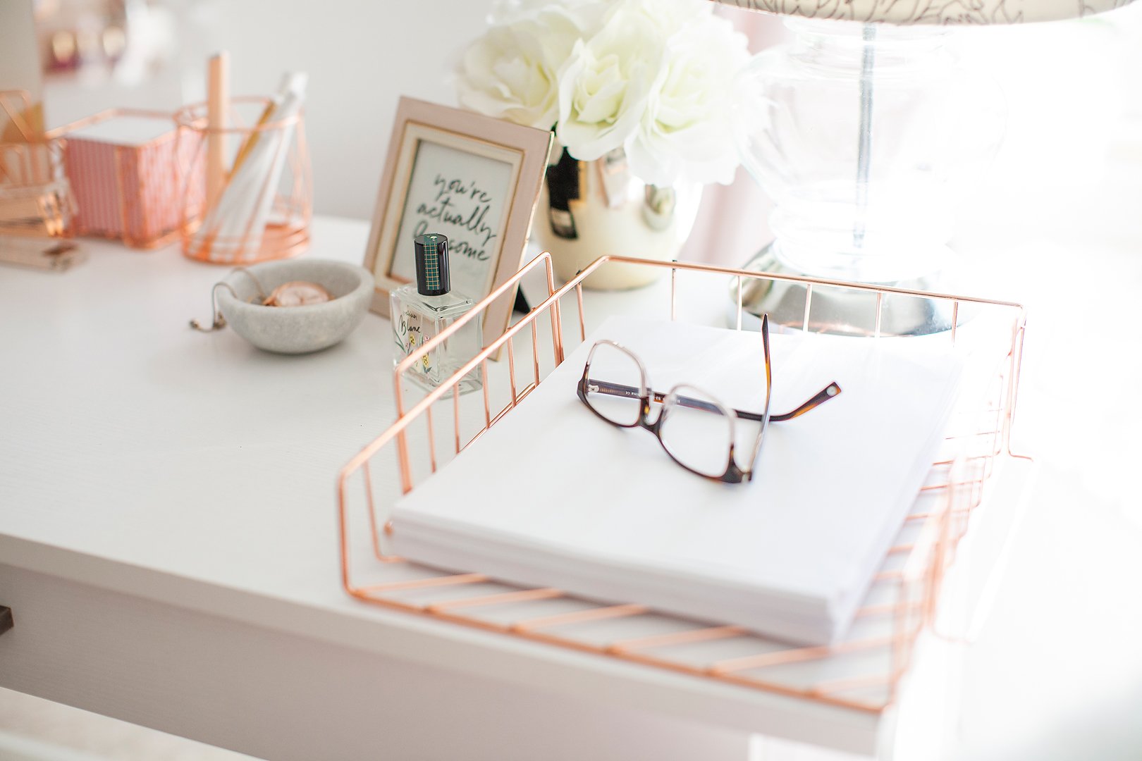 A bright, white desk with rose gold office supplies, including a file organizer with papers and glasses.