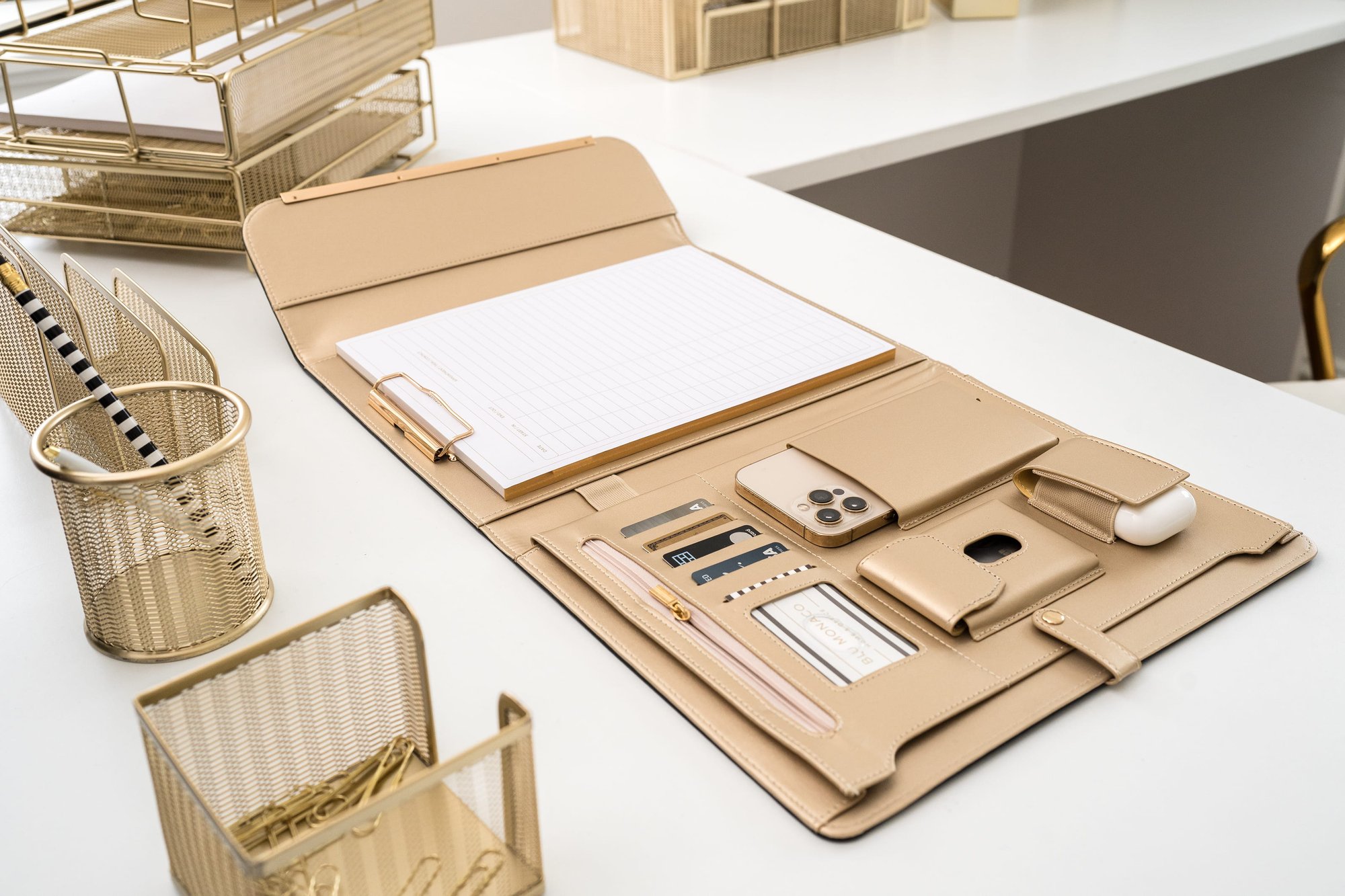 A beige desk organizer on a white desk, filled with a notepad, phone, and AirPods, surrounded by golden mesh office supplies.