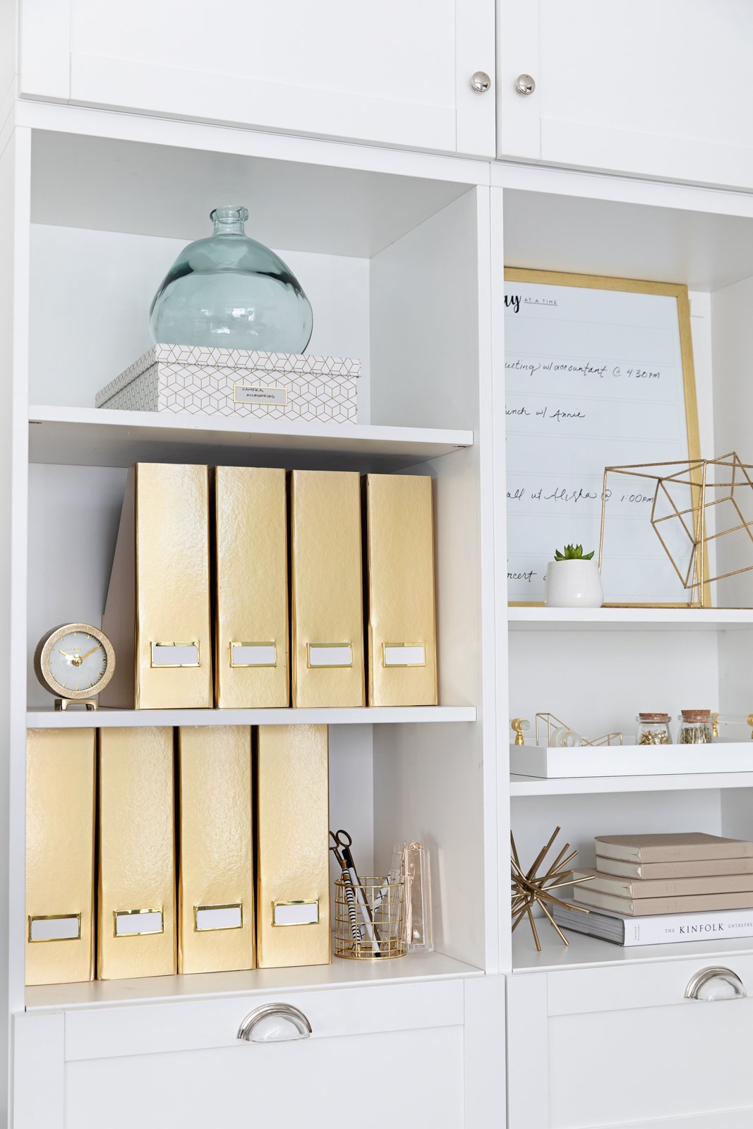 A neatly organized white home office bookshelf with gold binders, decor, and office supplies.