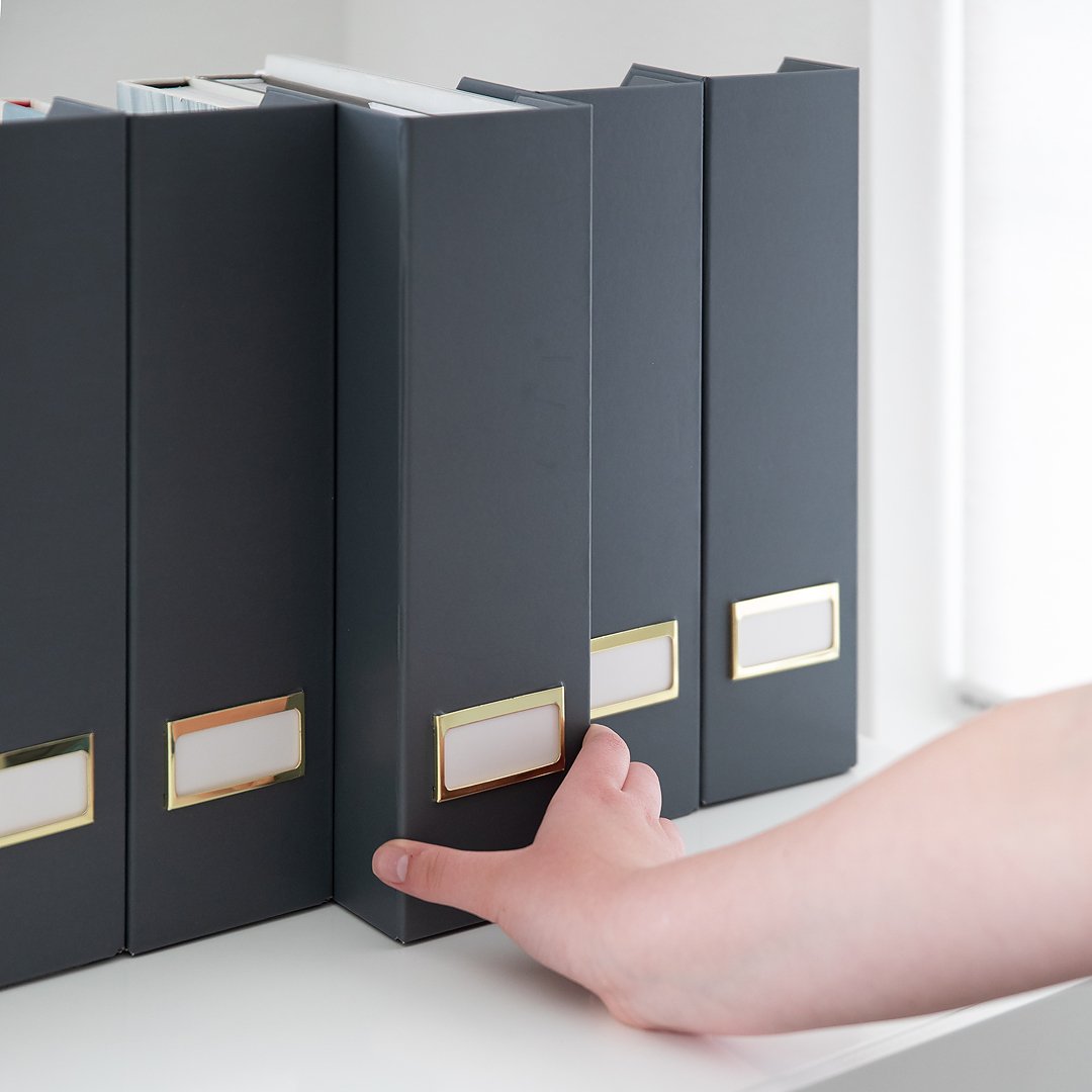 A person's hand reaches for a dark grey file box on a white shelf, among similar boxes.