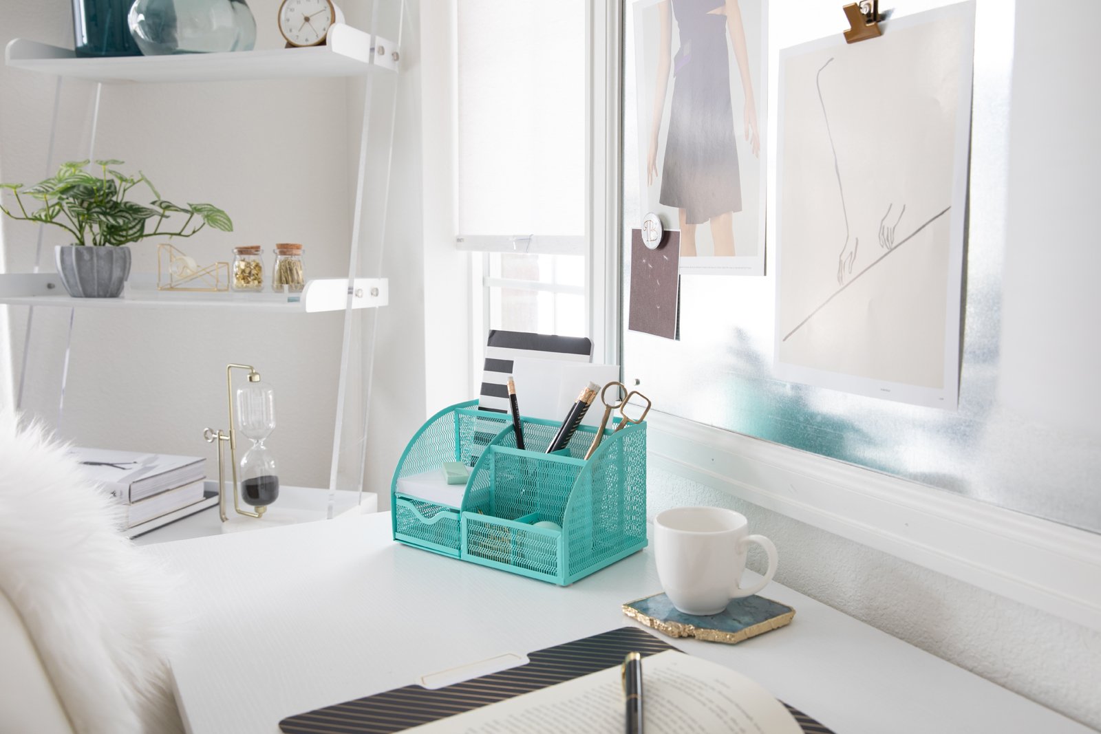 A well-lit white home office desk featuring a teal organizer, mug, notebook, and a white shelf.