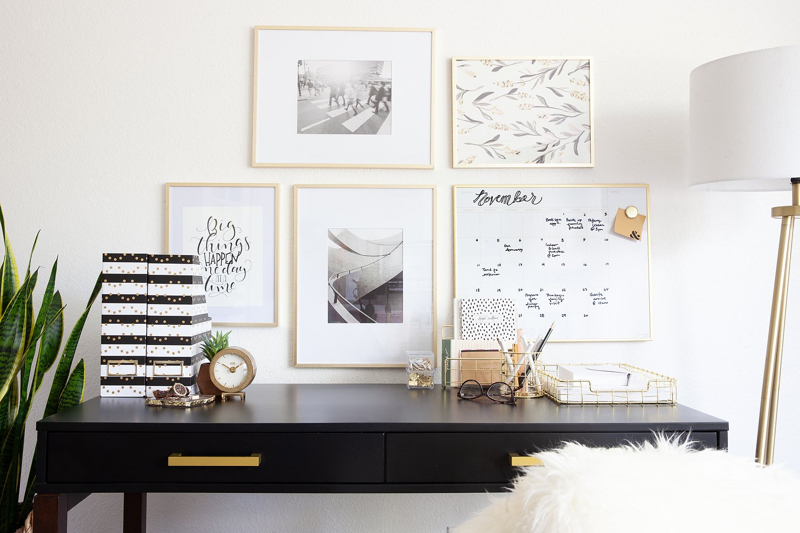 A stylish home office with a black desk, gold accessories, framed art, and a plant.