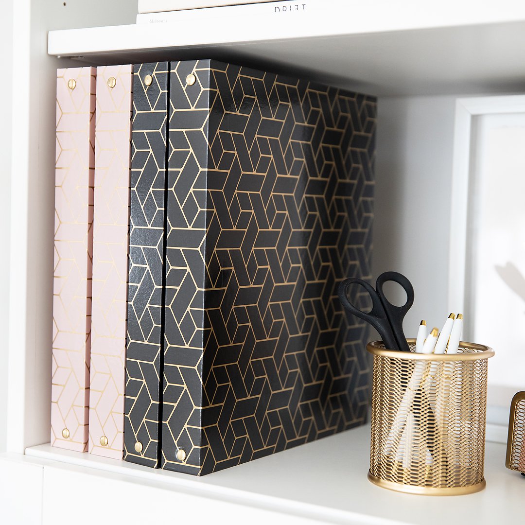 A neat white shelf holding stylish pink and black patterned binders, a gold pen holder with pens and black scissors.