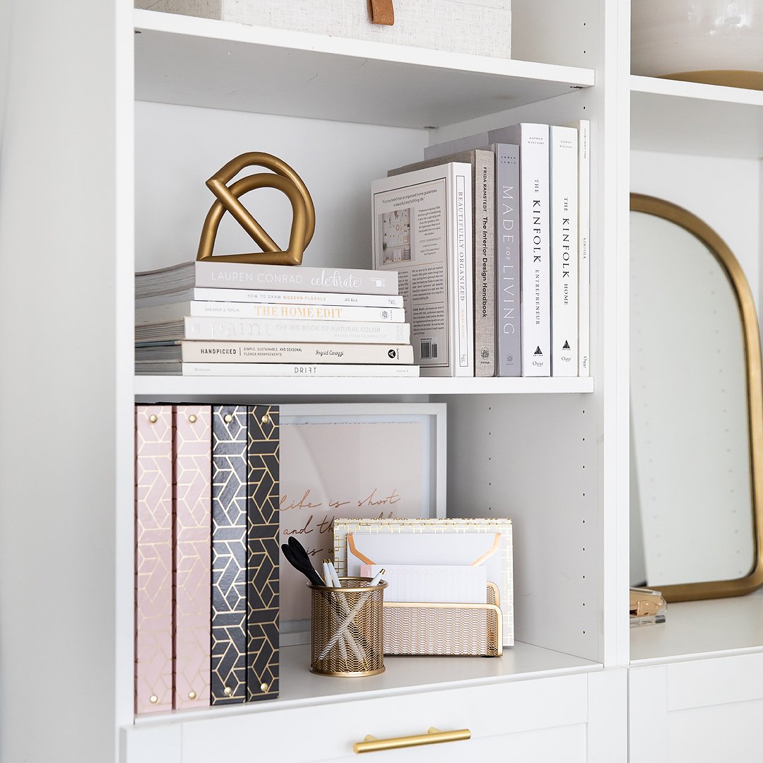 A beautifully organized white home office bookshelf with decorative items, books, binders, and gold office supplies.