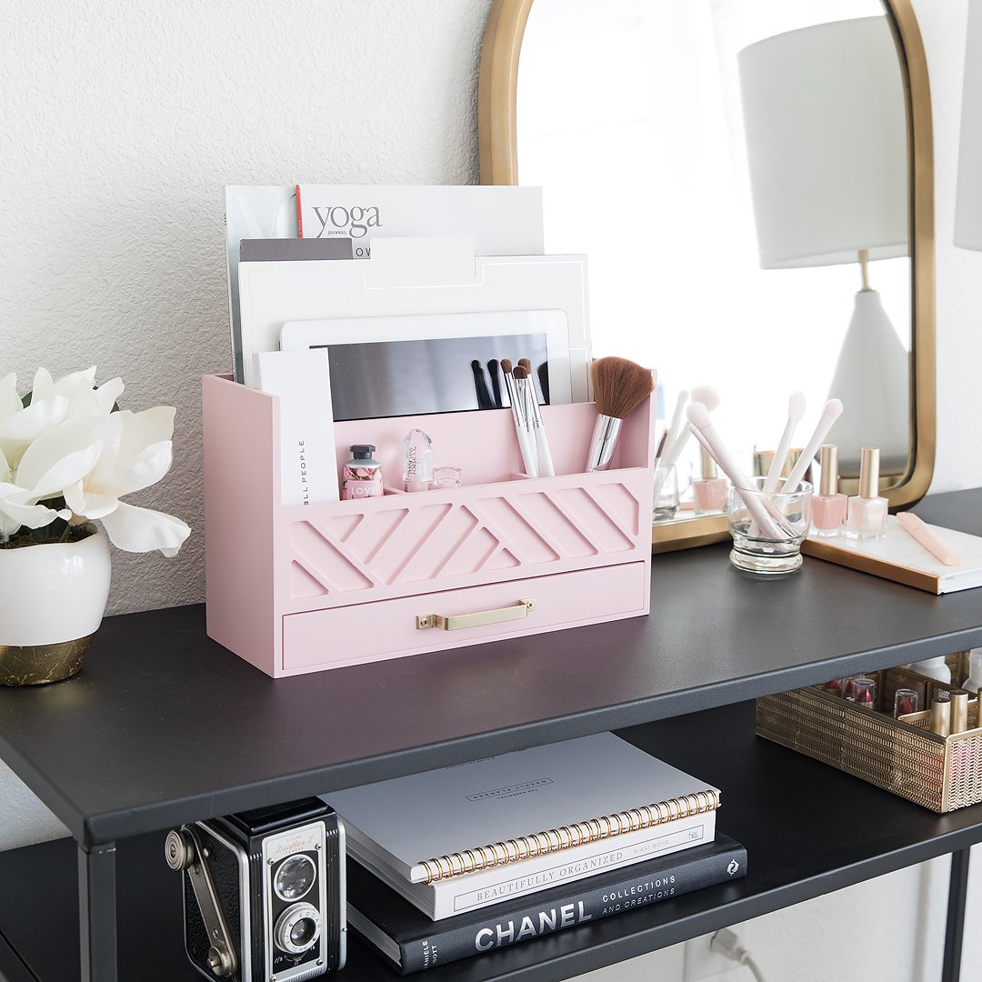 A pink desk organizer filled with magazines, makeup brushes, and cosmetics on a black desk.