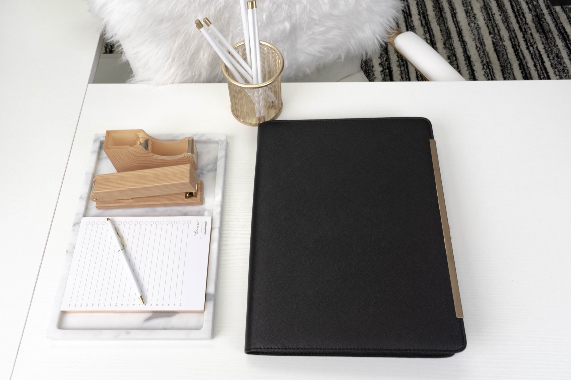 A top-down view of an organized white desk with a black folder, a marble tray with notepad and pen, a stapler, tape dispenser, and a gold pen holder with white pens.