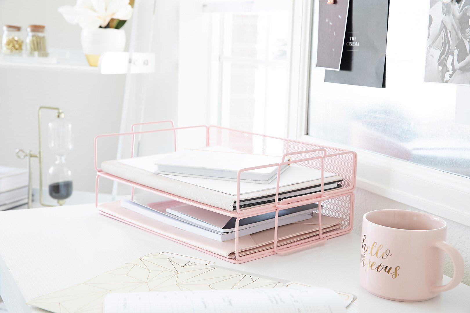 A neatly organized desk with a pink letter tray, white notebooks, a golden hourglass, and a pink mug.