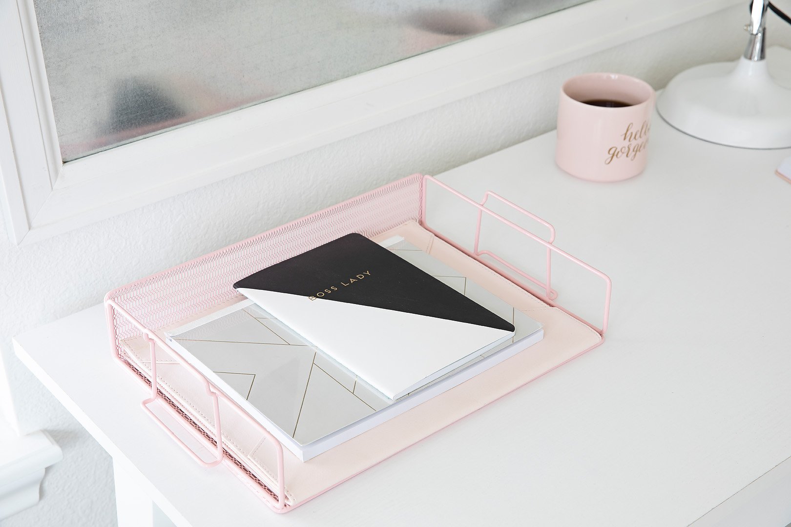 A pink mesh desk organizer holds notebooks, including one that says 'BOSS LADY', on a white desk.