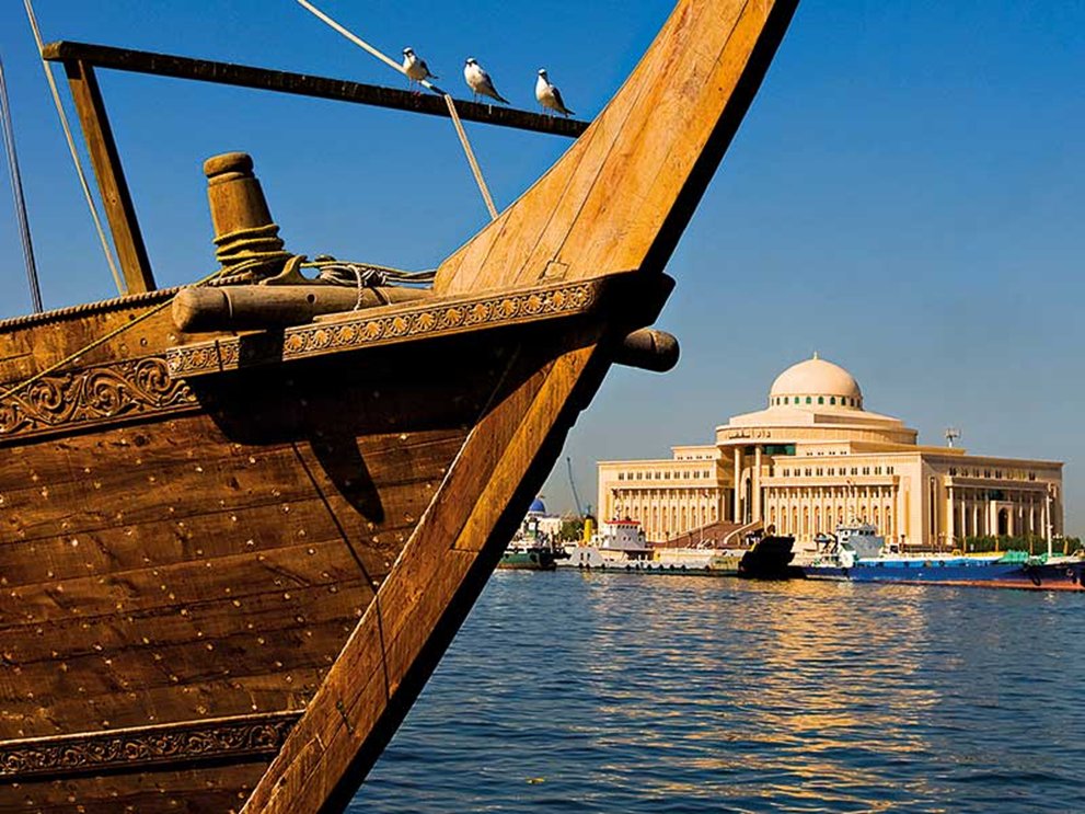 A traditional wooden Dhow boat's bow with birds, overlooking a grand domed building and harbor.