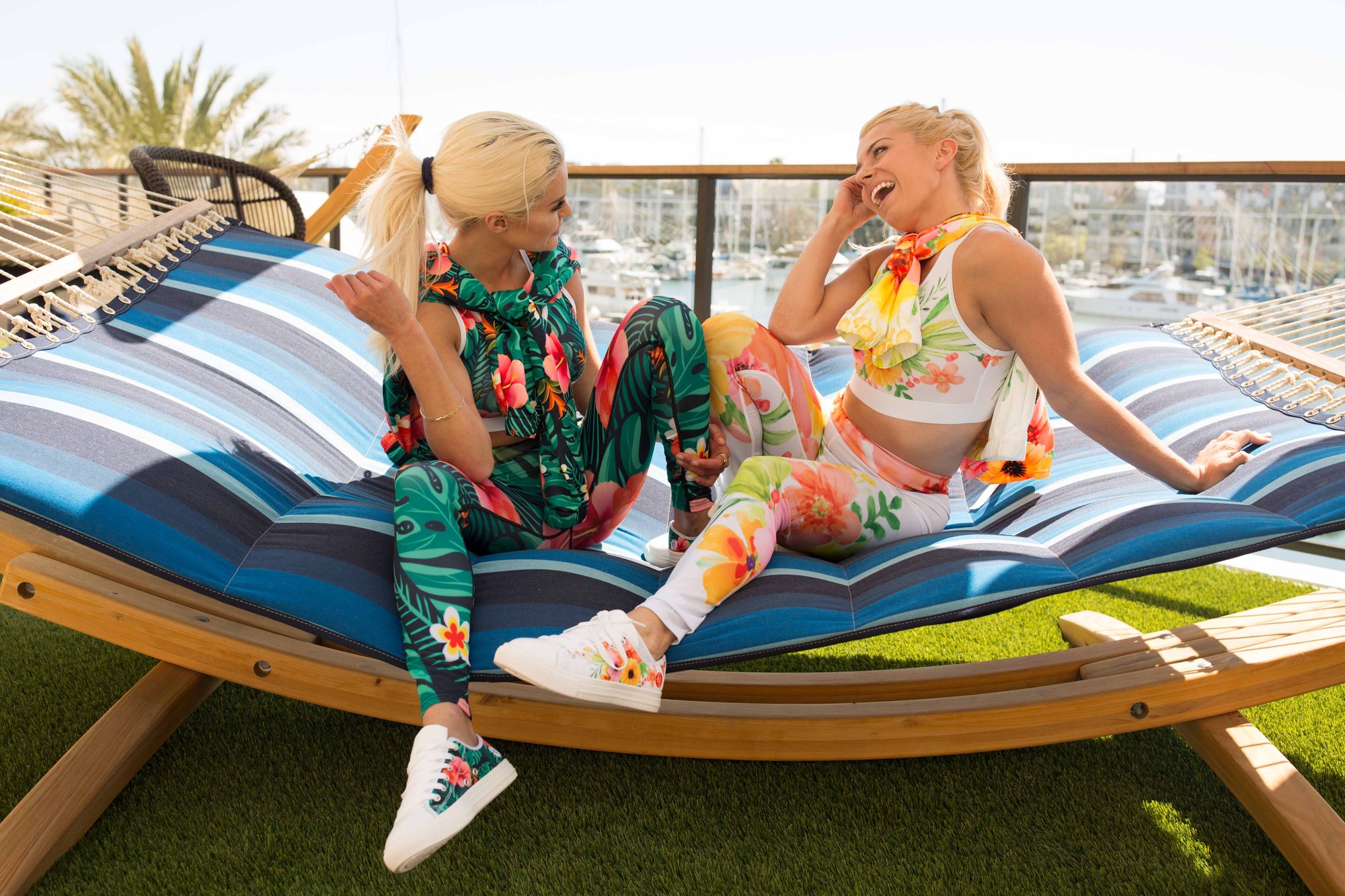 Two blonde women laughing in floral activewear, relaxing on a striped hammock with a harbor view.