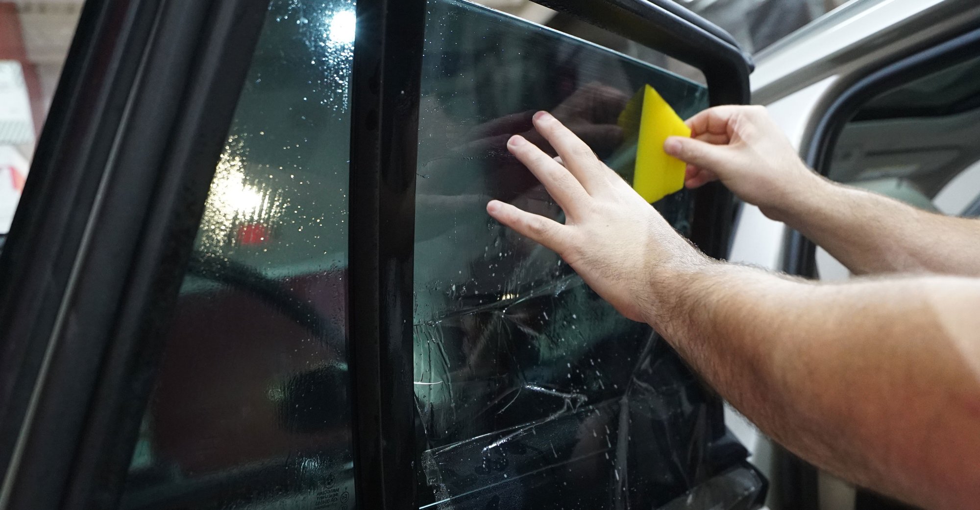 Close-up of hands applying tint film to a car window with a yellow squeegee.