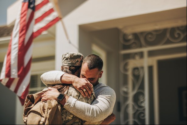 A military service member in uniform is embraced by a man in front of a house with an American flag.