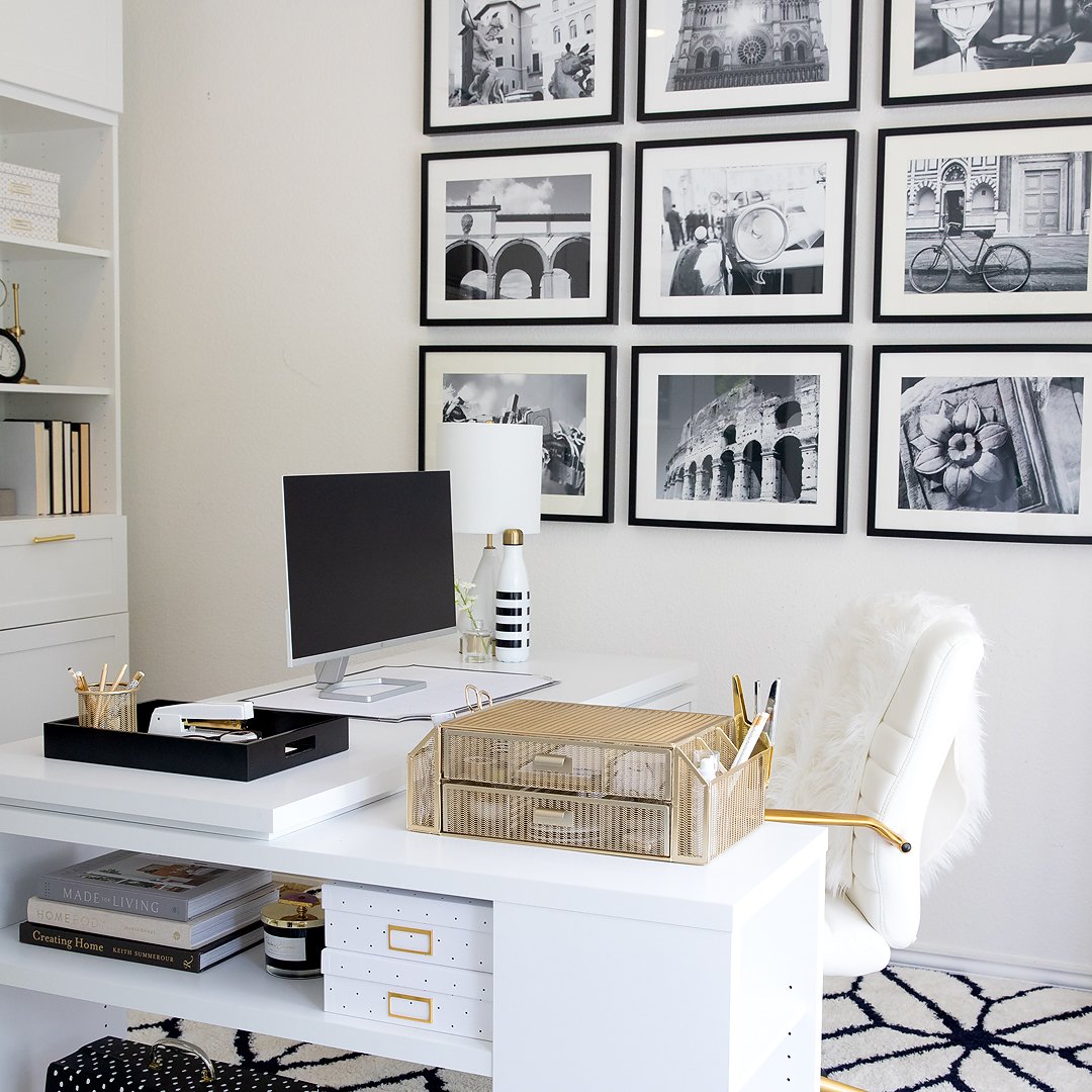 A stylish and organized white home office desk with a computer, gold decor, and black and white framed photos.