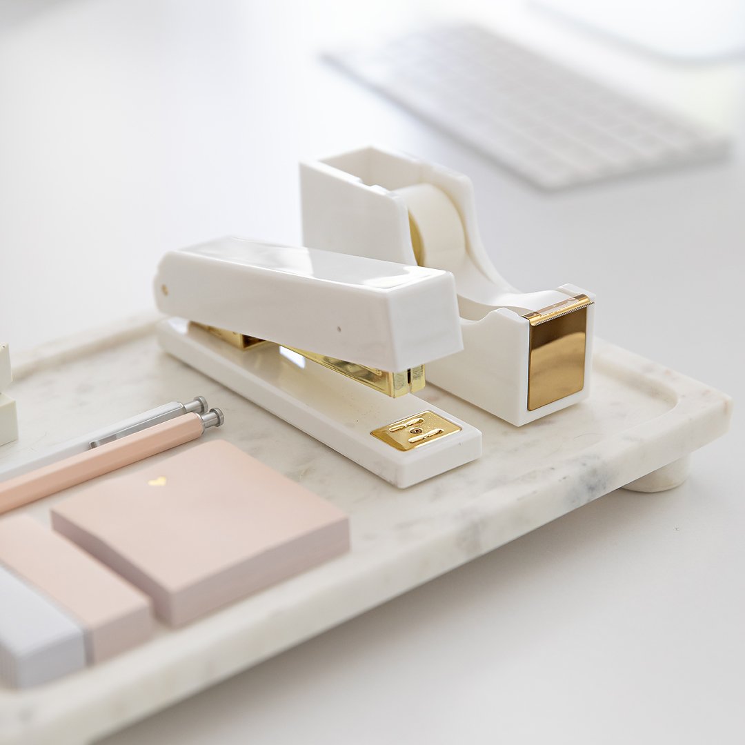 Elegant white and gold stapler, tape dispenser, and pink sticky notes on a marble desk tray.