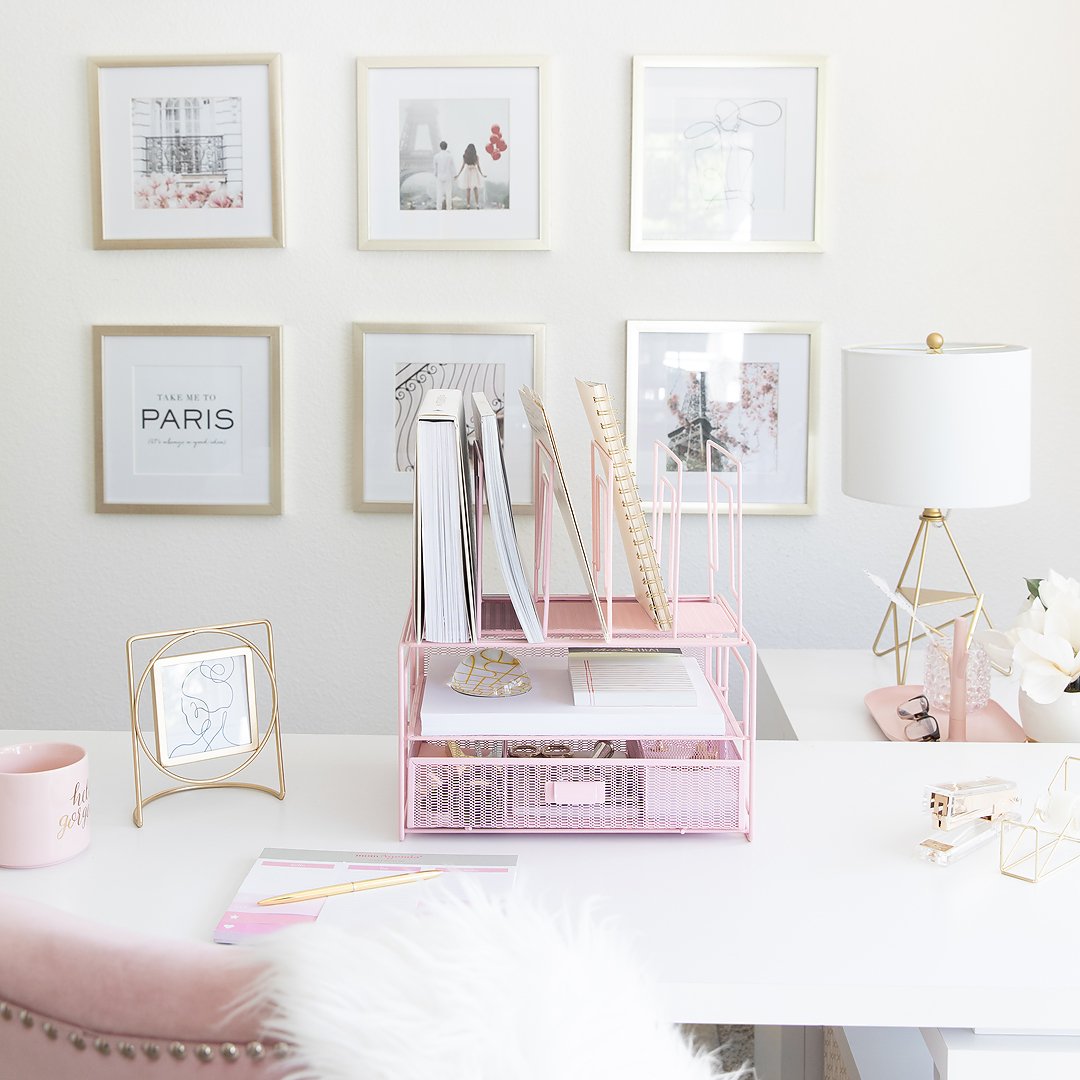 A stylish pink and gold home office desk with organized supplies, framed art, and a lamp.
