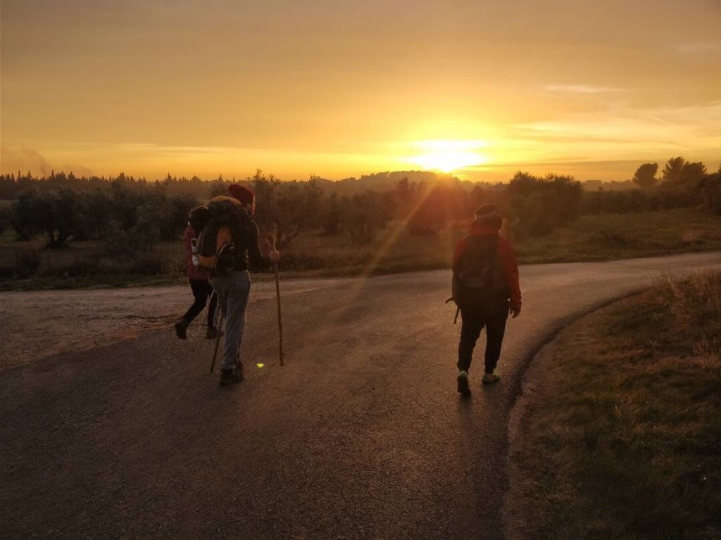 Trois randonneurs avec des sacs à dos et bâtons marchent sur une route au lever du soleil.
