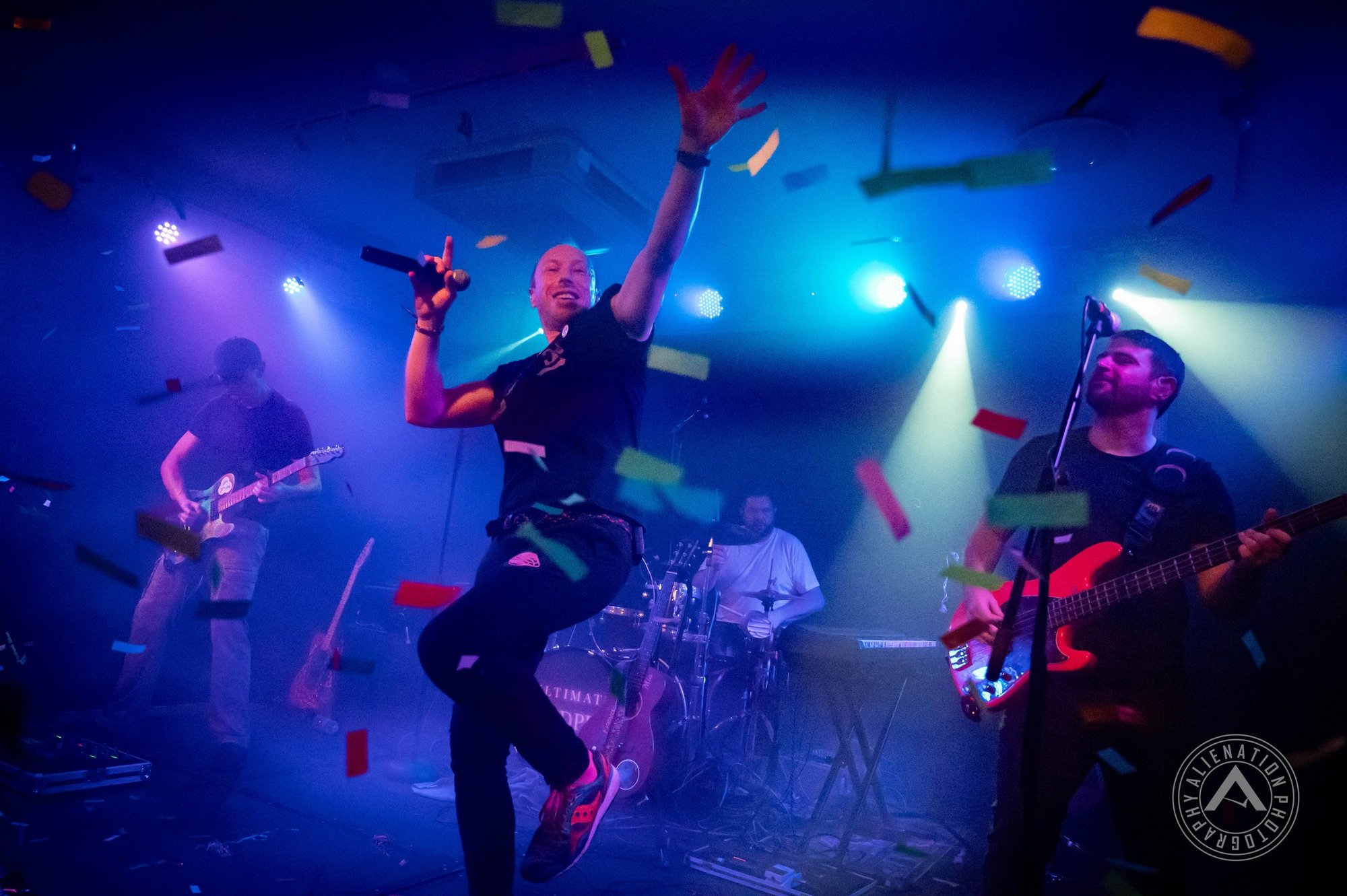 A band performs on a stage under vibrant blue lights with confetti falling, featuring an energetic singer.
