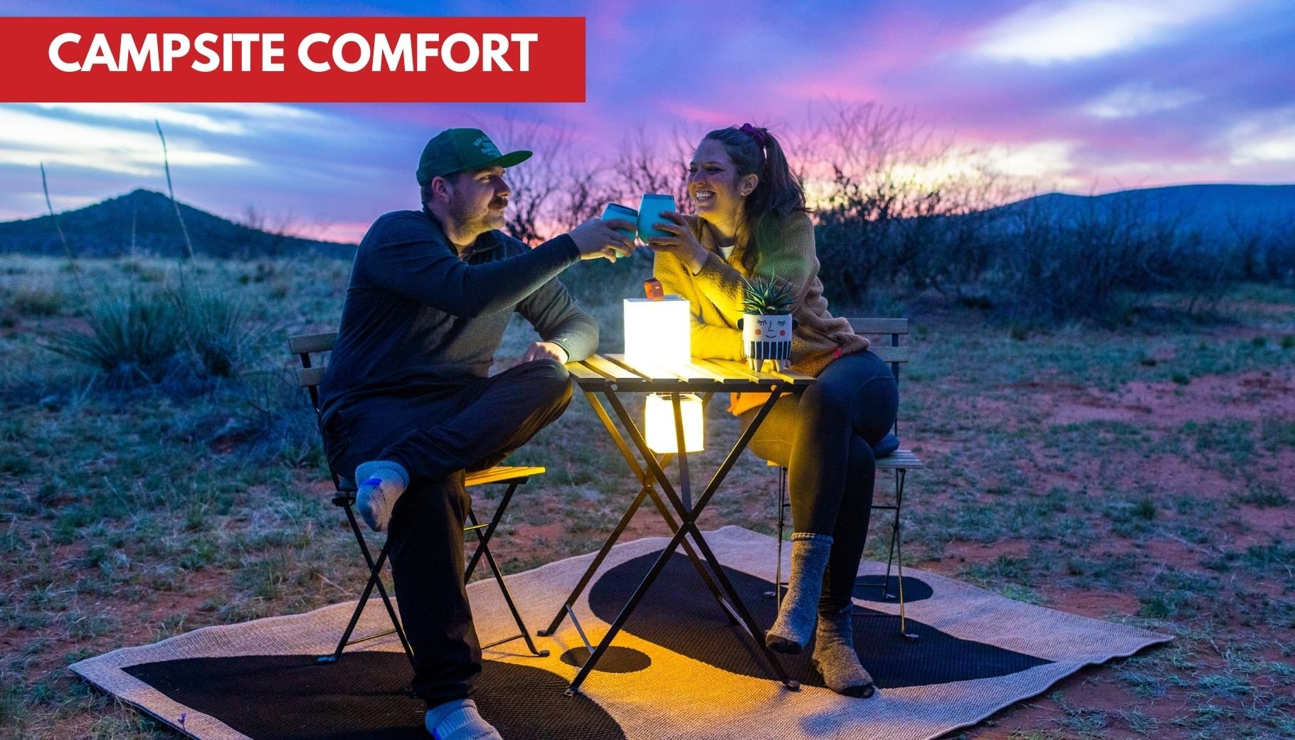 A couple toasts with drinks at a campsite table, illuminated by LuminAID lanterns under a colorful sunset.