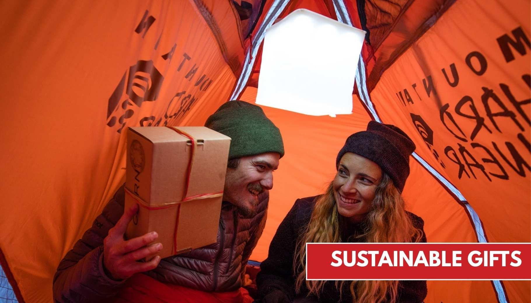 A man holds a gift box and smiles at a woman inside an orange tent with a bright LuminAID lantern hanging from the tent ceiling.