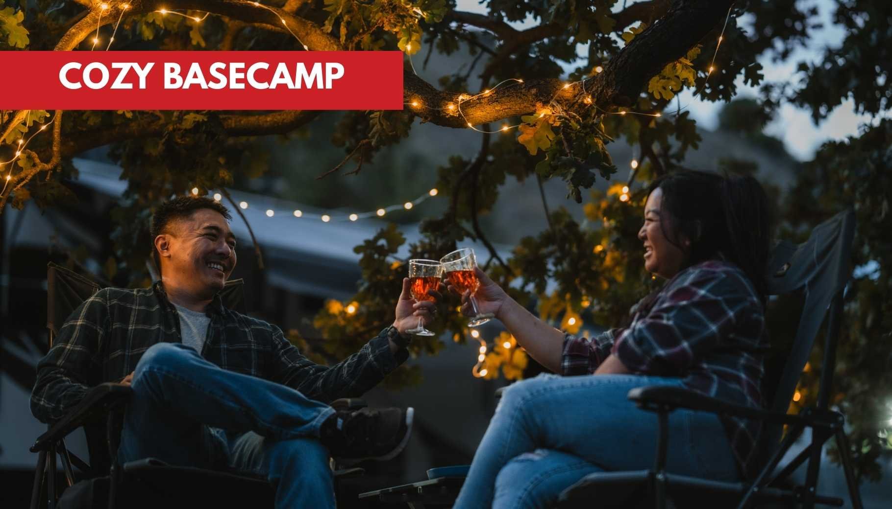 A happy couple toasts with drinks in camping chairs under festive string lights at dusk.