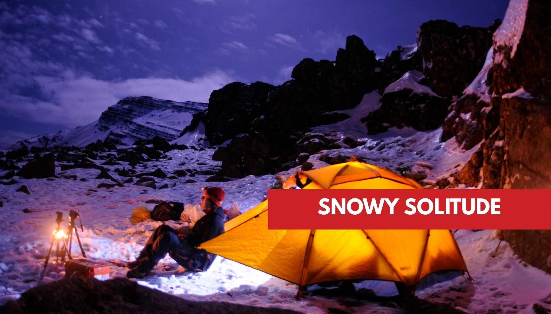 A person camping in snowy mountains at night, illuminated by a tent and small light.