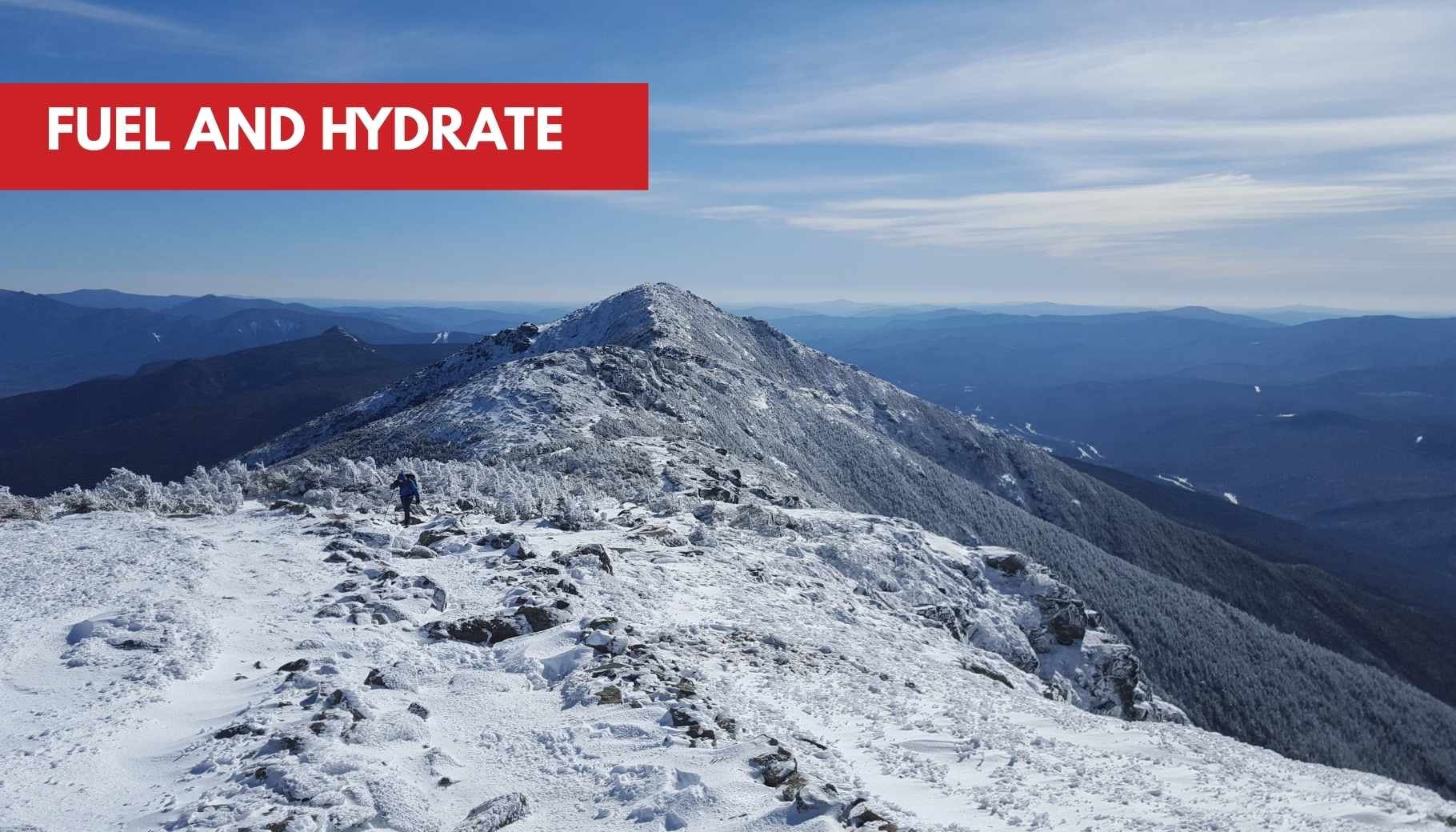 A hiker on a snow-covered mountain ridge under a bright blue sky, with distant peaks.