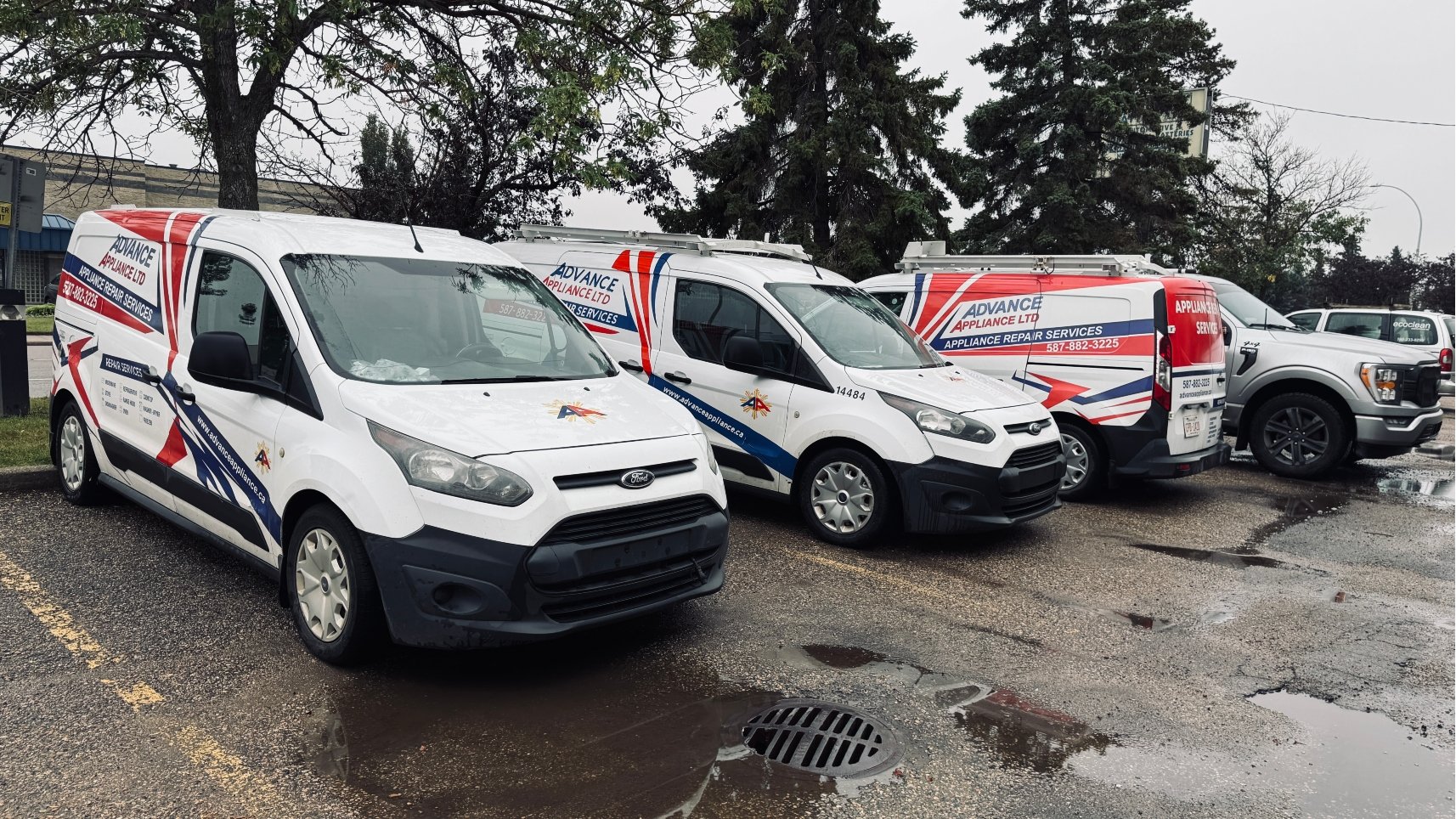 Several white service vans and a pickup truck from Advance Appliance Ltd parked on a wet lot.