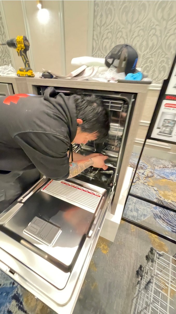 A person in a black hoodie bends over, meticulously installing a new stainless steel dishwasher appliance.