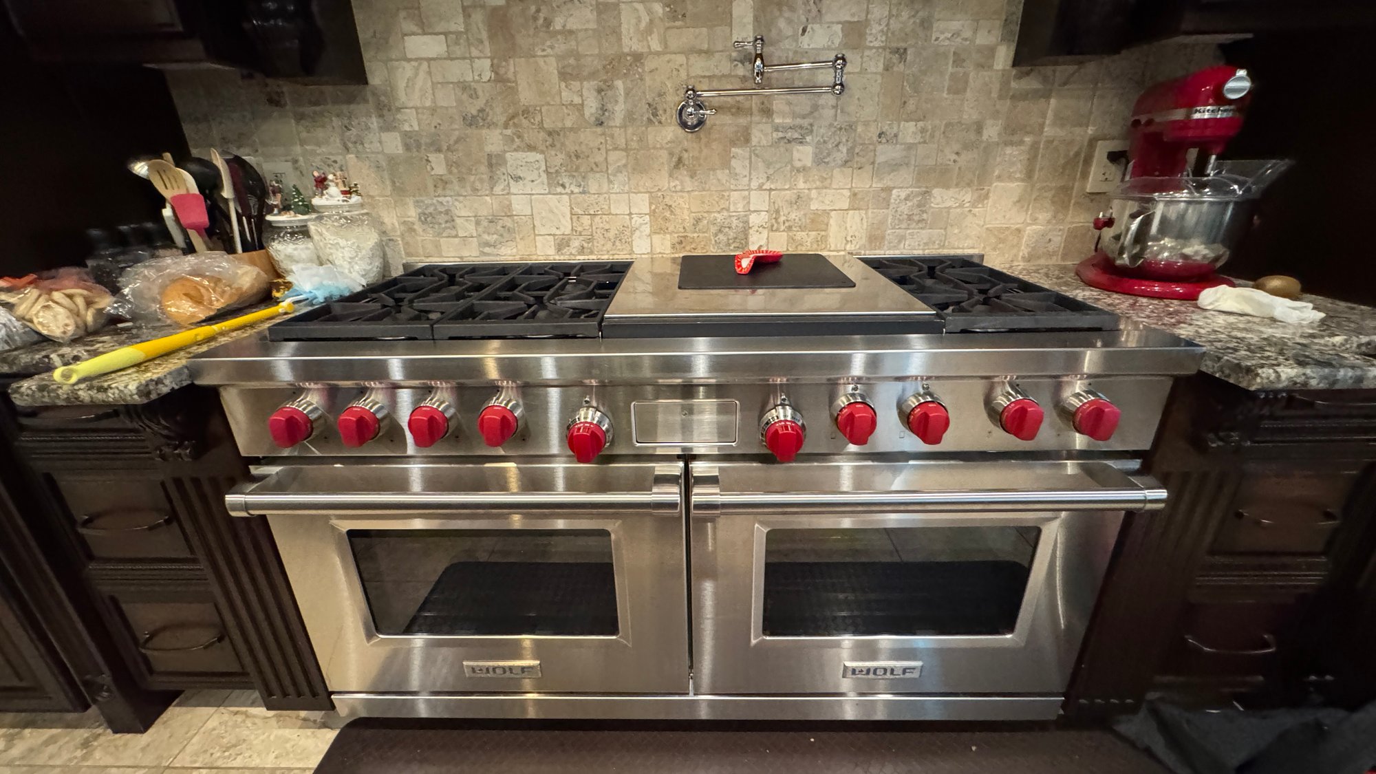 A high-end stainless steel Wolf range with red knobs, dual ovens, and a pot filler above a tiled backsplash.