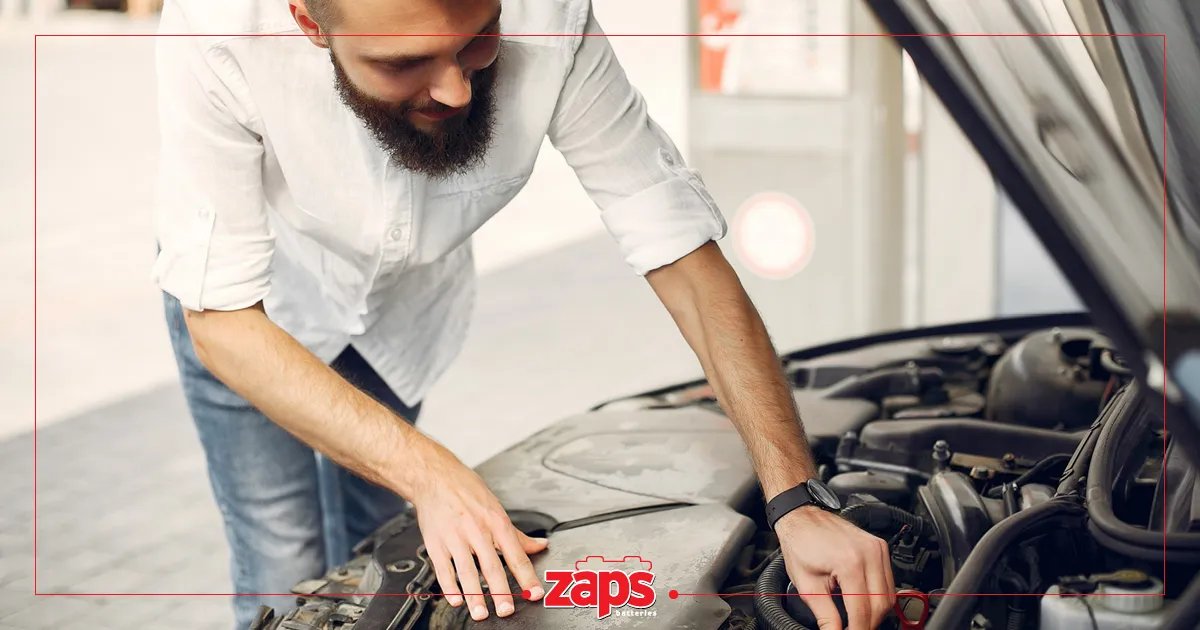 Άντρας με γενειάδα, φορώντας λευκό πουκάμισο, επισκευάζει το αυτοκίνητό του, κοιτάζοντας κάτω από το καπό.