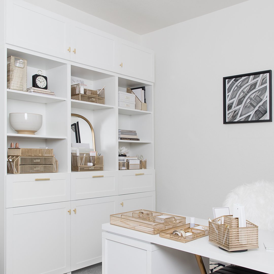 A bright, organized home office with white built-in shelves and a desk with gold accessories.