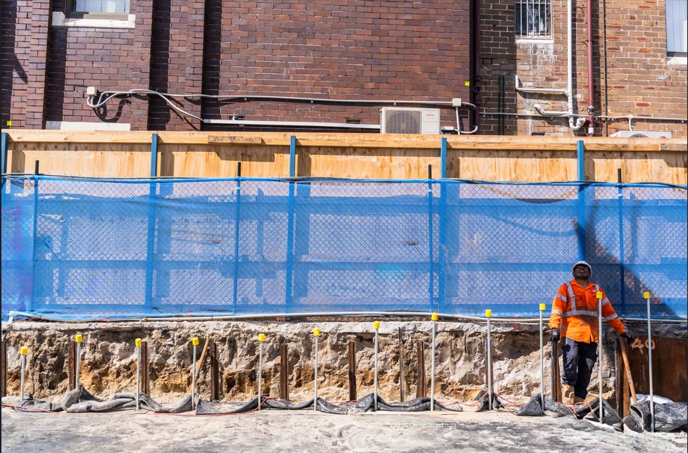 A construction worker in an orange vest and hard hat stands in an excavated trench.