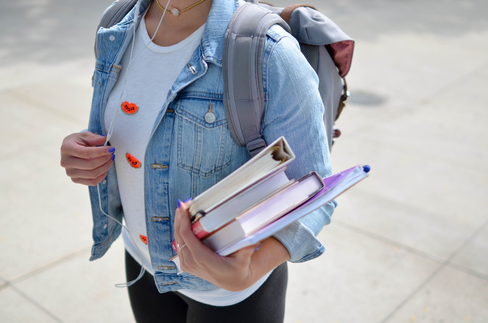 A student in a denim jacket and backpack holds books while listening to white headphones.