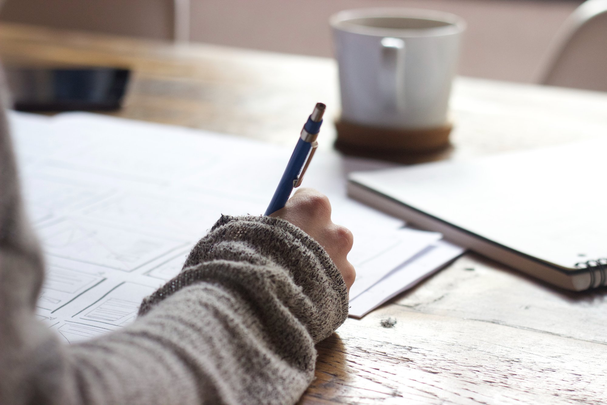 A person's hand in a sweater holds a blue pen, drawing on paper next to a coffee mug on a wooden table.