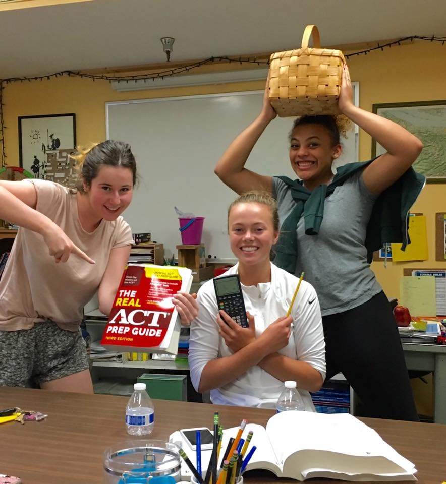 Three smiling young women in a classroom, two holding study materials and one a basket.