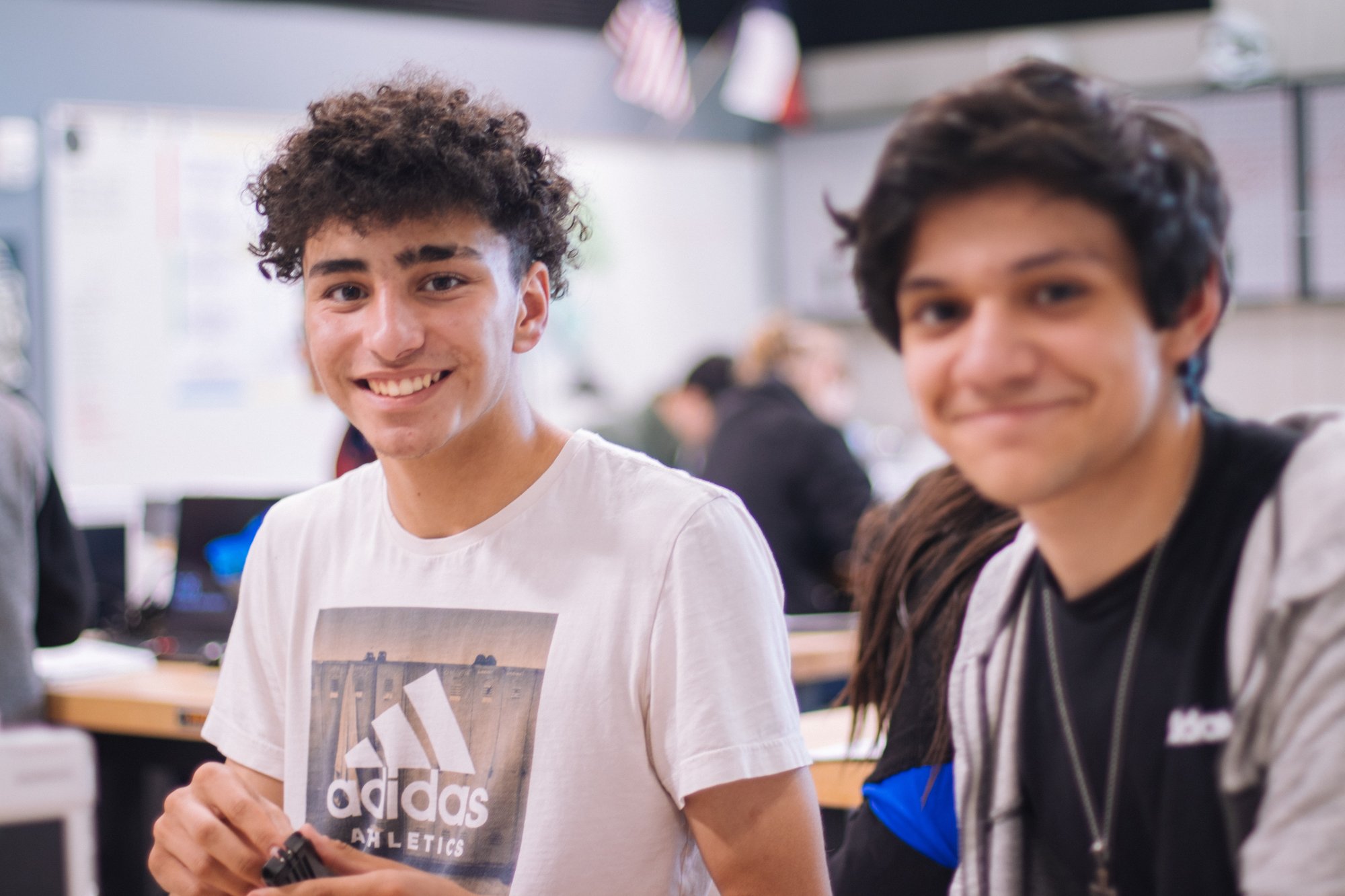 Two smiling male students in a classroom, one wearing an Adidas t-shirt and holding an object.