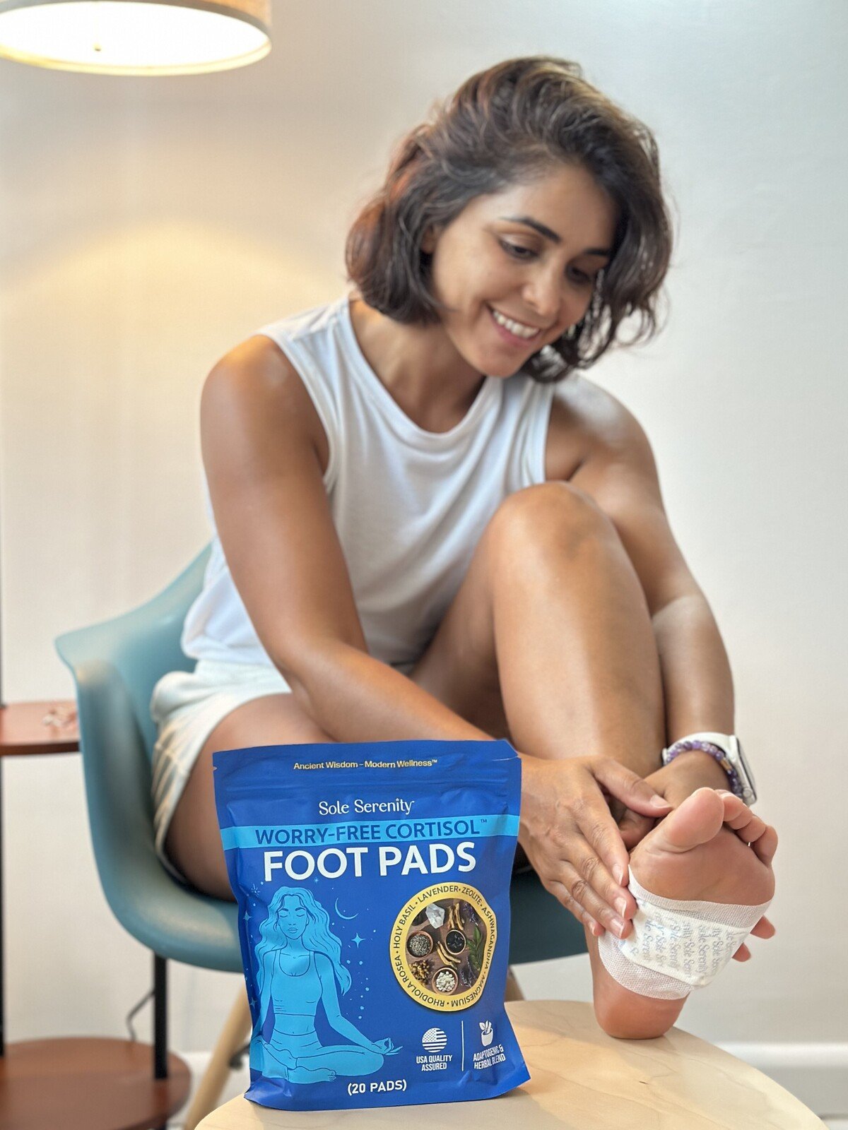 A smiling woman applies a cortisol-reducing foot pad to her sole, with the product package nearby.