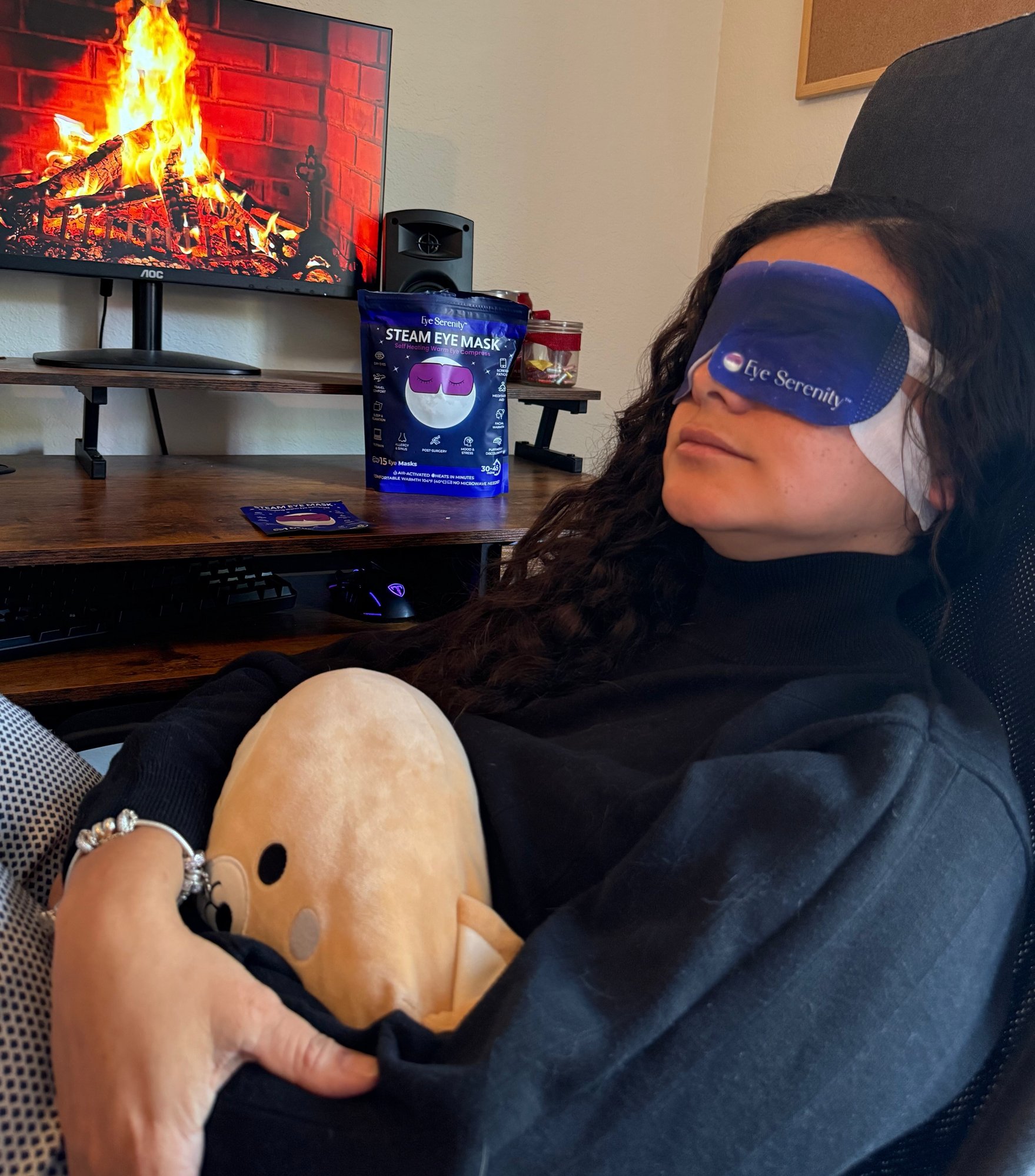 Woman wearing a steam eye mask and holding a plush toy, relaxing by a fireplace video.