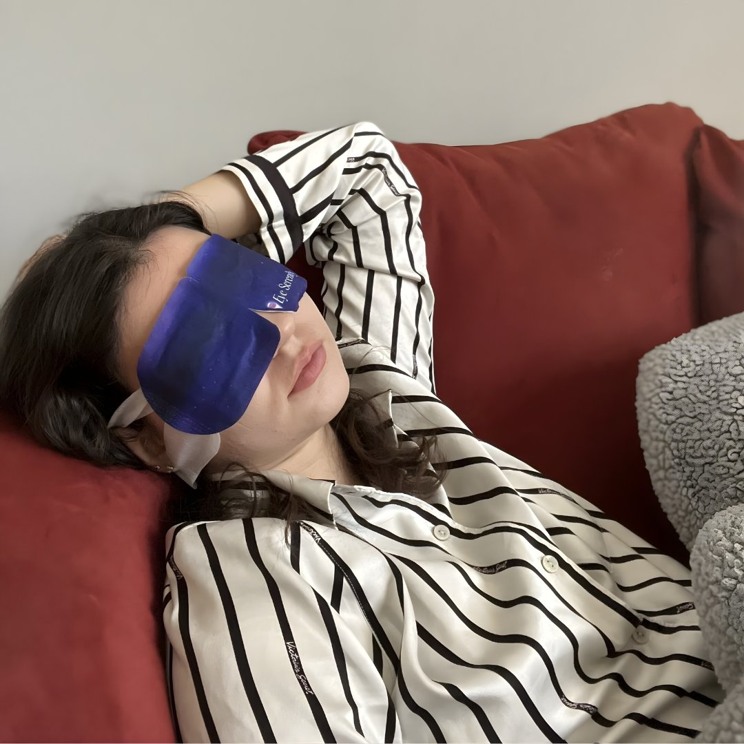 A person in striped pajamas with a blue eye mask relaxing on a red couch.
