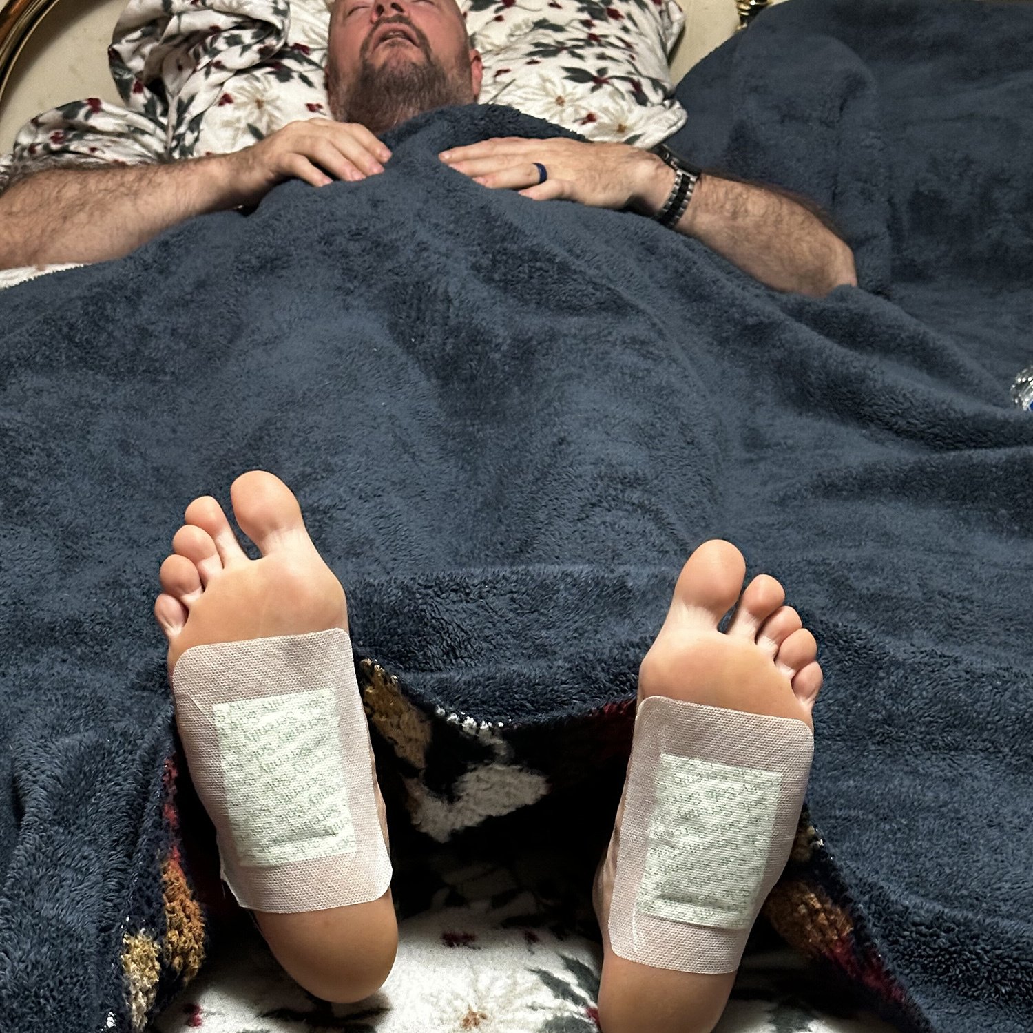 A person rests in bed with detox foot pads applied to the soles of both feet.