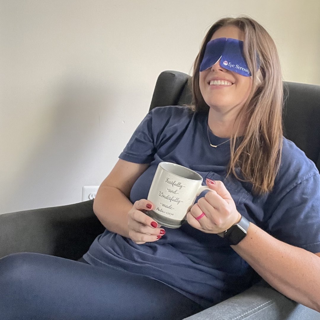 A woman smiles while relaxing in a chair, wearing an eye mask and holding a mug.