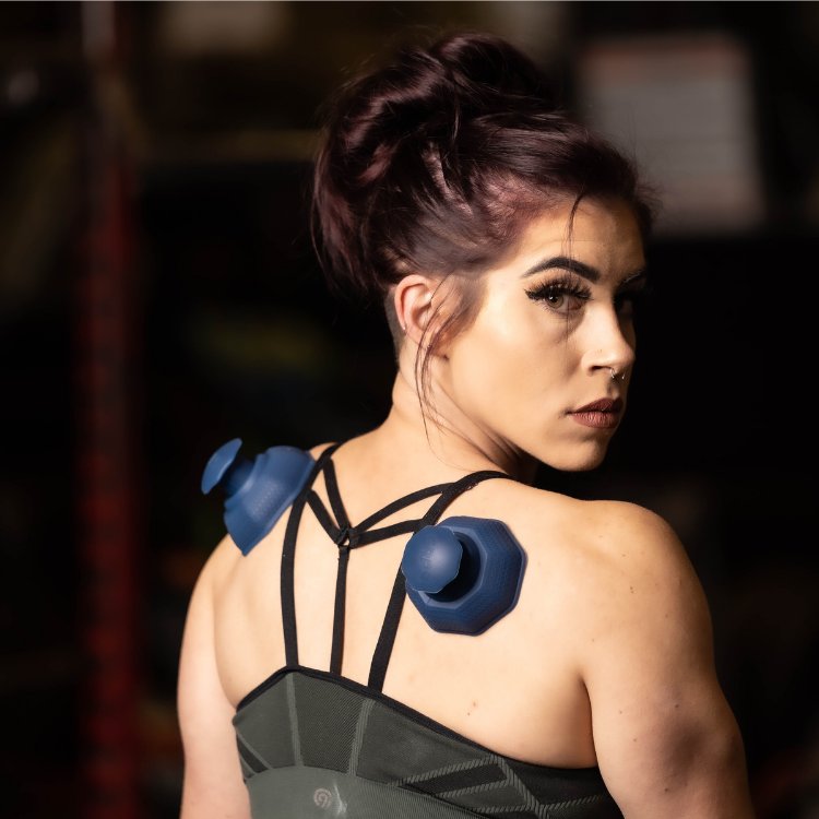 A woman with a dark sports bra wears two blue muscle therapy devices on her shoulders, looking over her left shoulder.