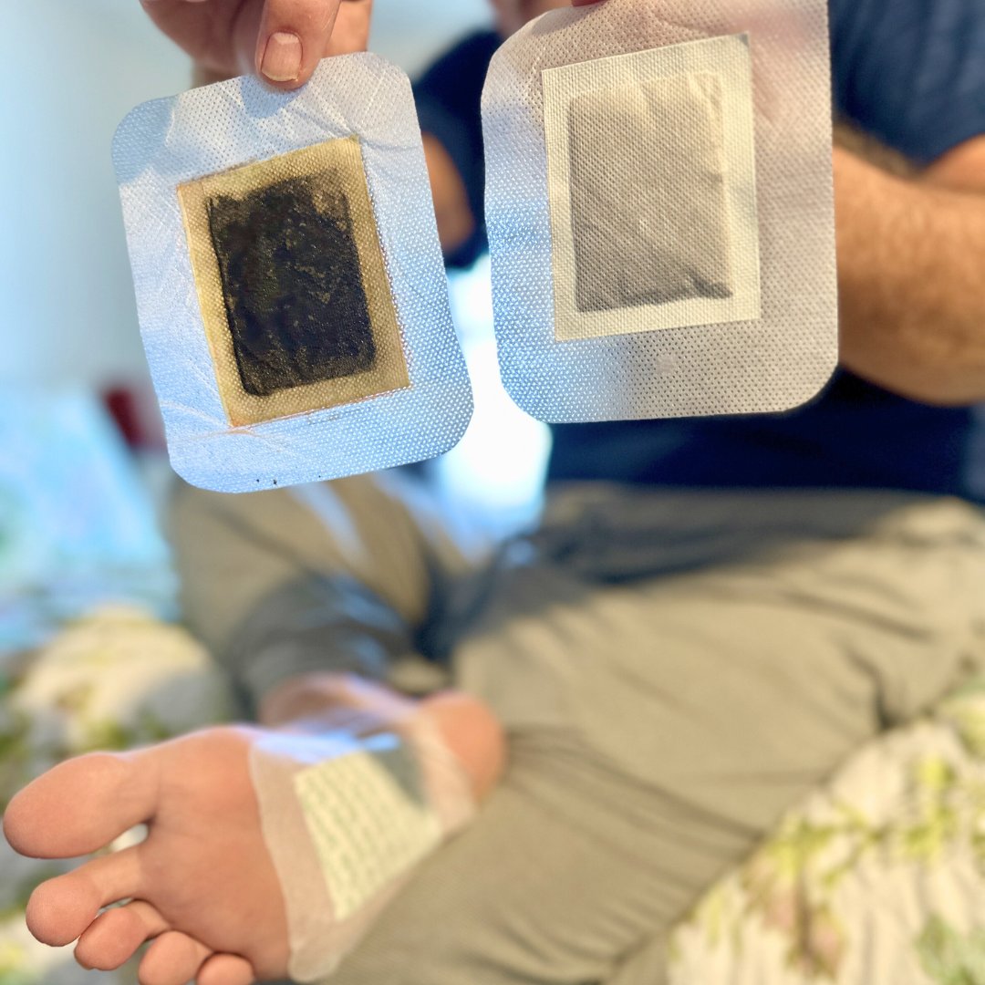 A person shows a dirty and clean detox foot pad, with another pad on their foot.