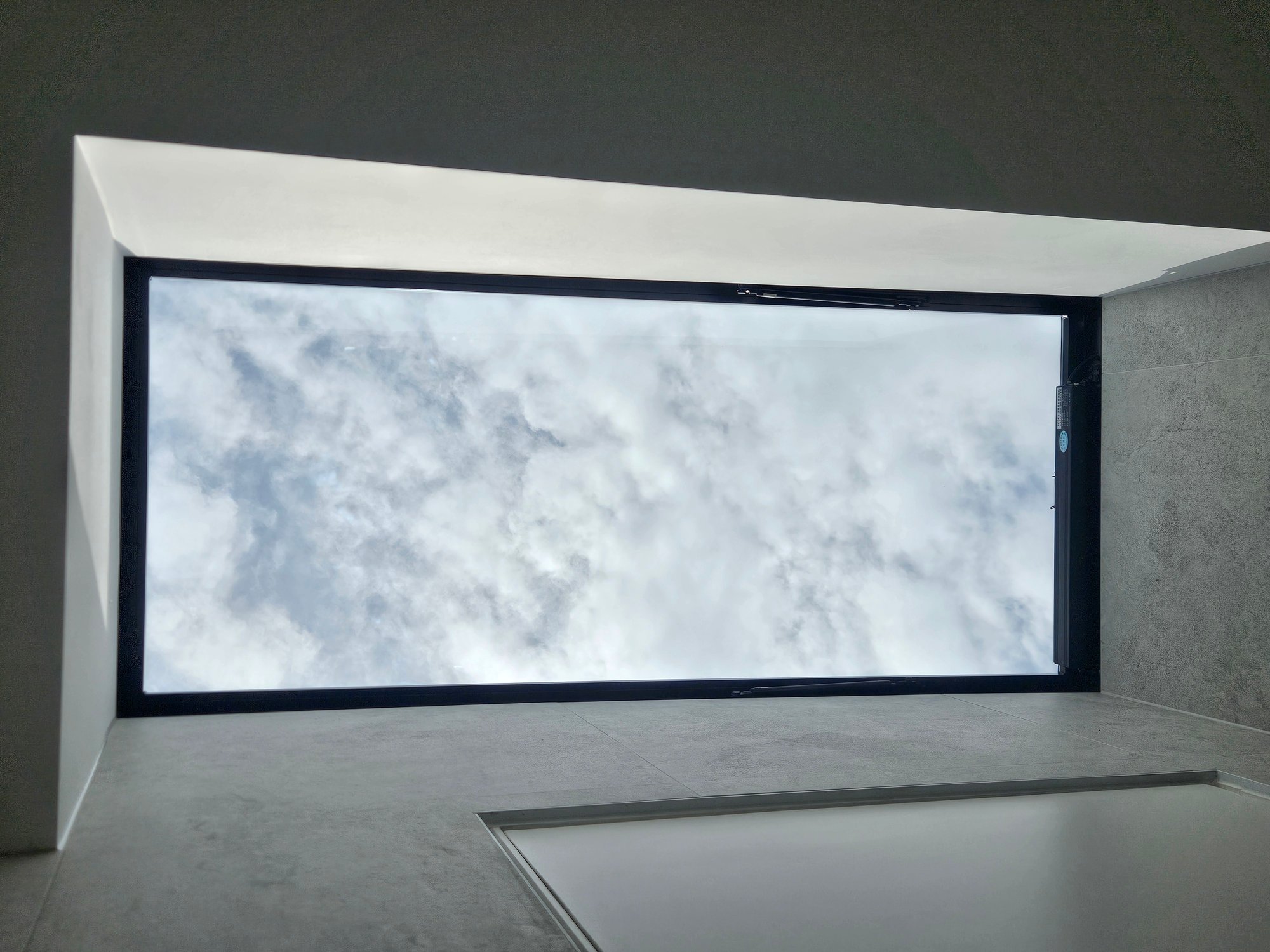 View from below a rectangular skylight showing a cloudy sky and surrounding grey interior walls.