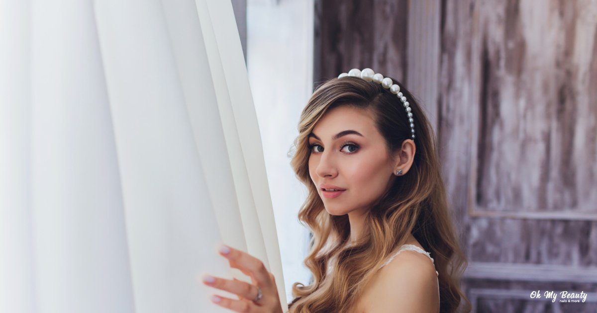 Ρομαντικό Νυφικό Μακιγιάζ: Ο Απόλυτος Οδηγός για το 2026 Κοντινό πλάνο μιας γυναίκας με μακιγιάζ, κυματιστά μαλλιά και στέκα με πέρλες, κοιτάζει ανάμεσα σε λευκές κουρτίνες.