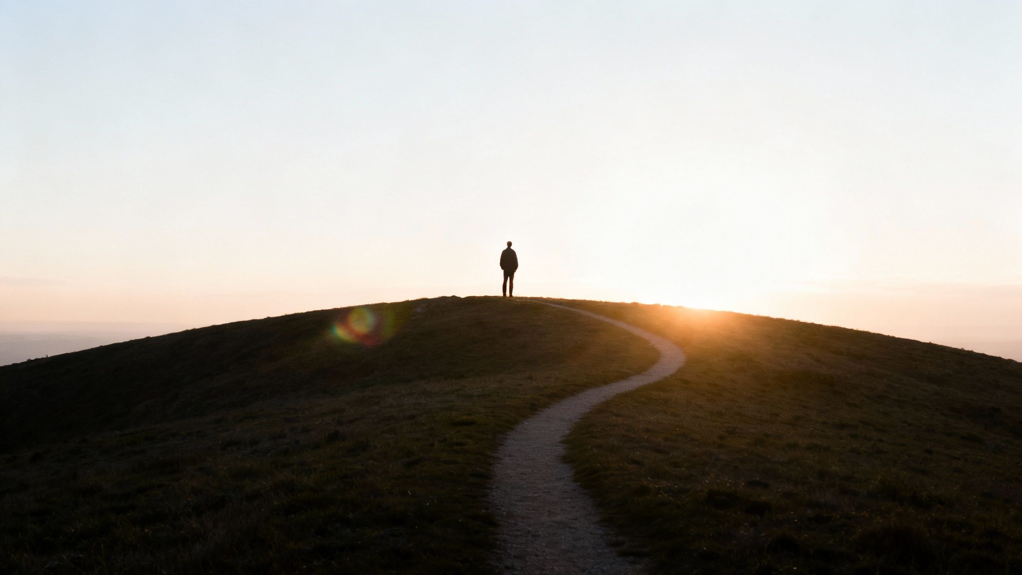 A lone figure stands on a grassy hill, gazing at a vibrant sunset over a winding path.